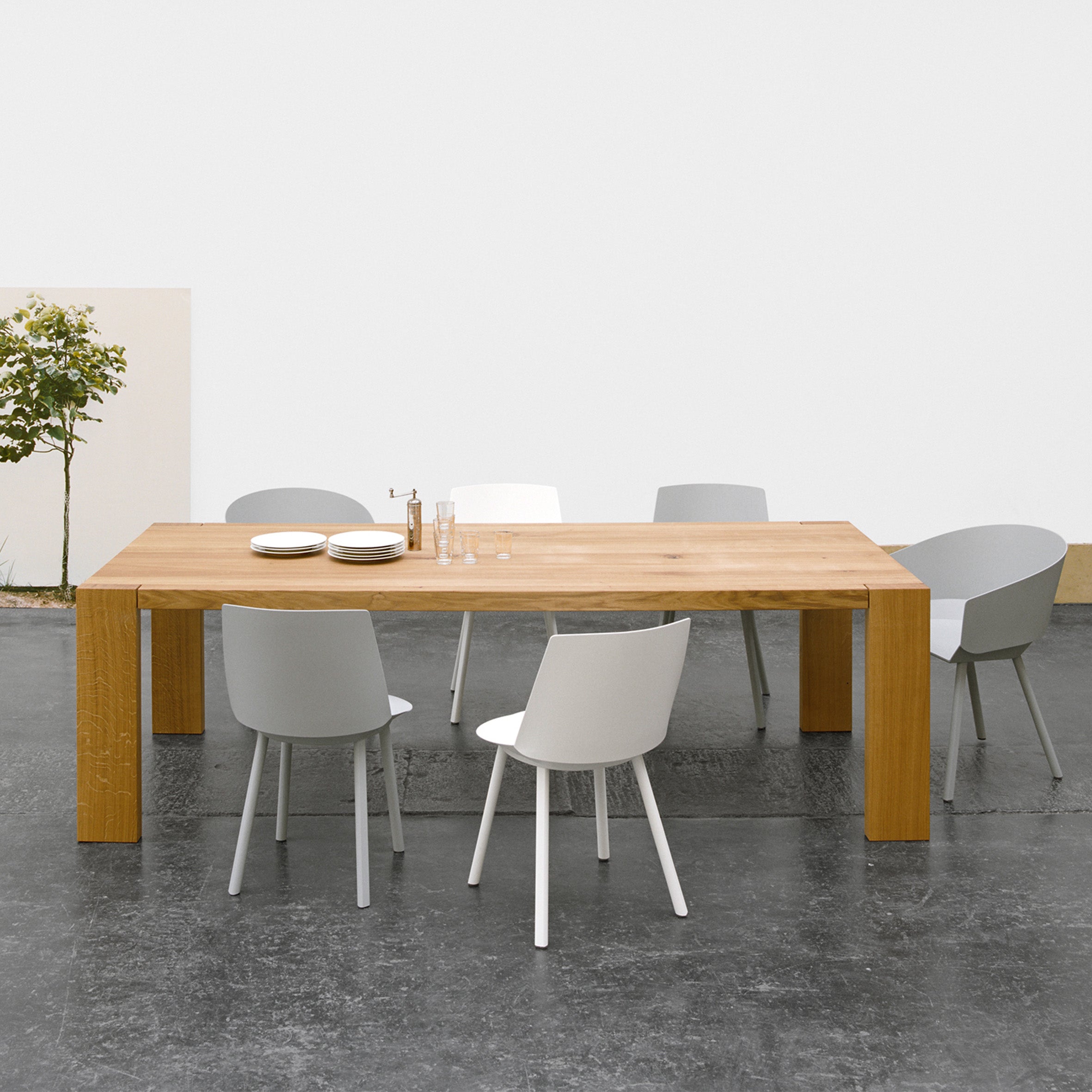 In a minimalist dining area, the E15 TA17 LONDON solid wood table is set for three, surrounded by white chairs. Plates and glasses adorn the table, while a small potted plant rests nearby against a plain wall with a casually propped tree picture.