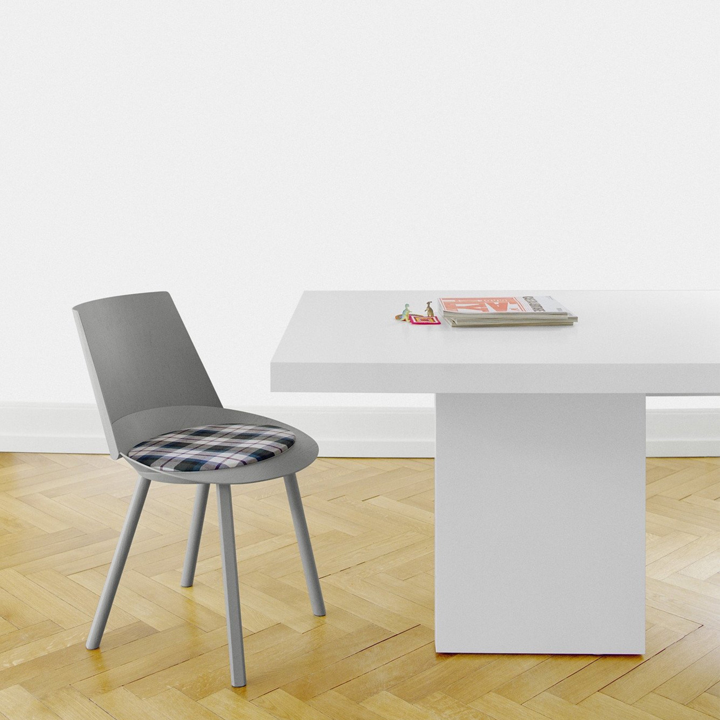 A modern, minimalist room showcases a white E15 TA18 ZEHN table with a sturdy pedestal base, adorned with two books and glasses. Beside it stands a gray chair with a plaid cushion on light wood parquet flooring, enhancing the contemporary European oak vibe.