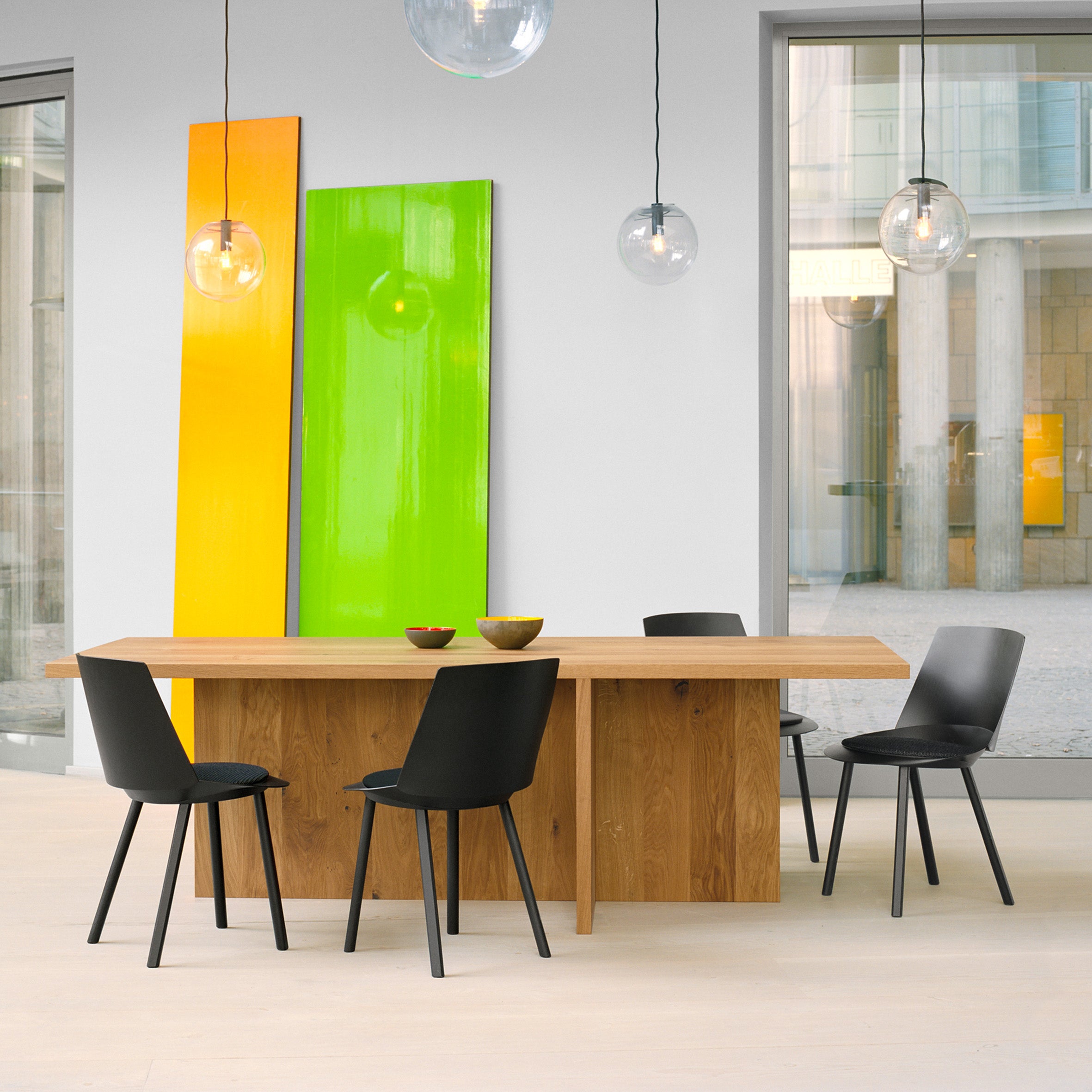 The minimalist dining room showcases the TA18 ZEHN solid wood table by E15, crafted from European oak, accompanied by four black chairs and three hanging glass pendant lights. Bright yellow and green panels energize the space, with large windows providing ample light and a view beyond.