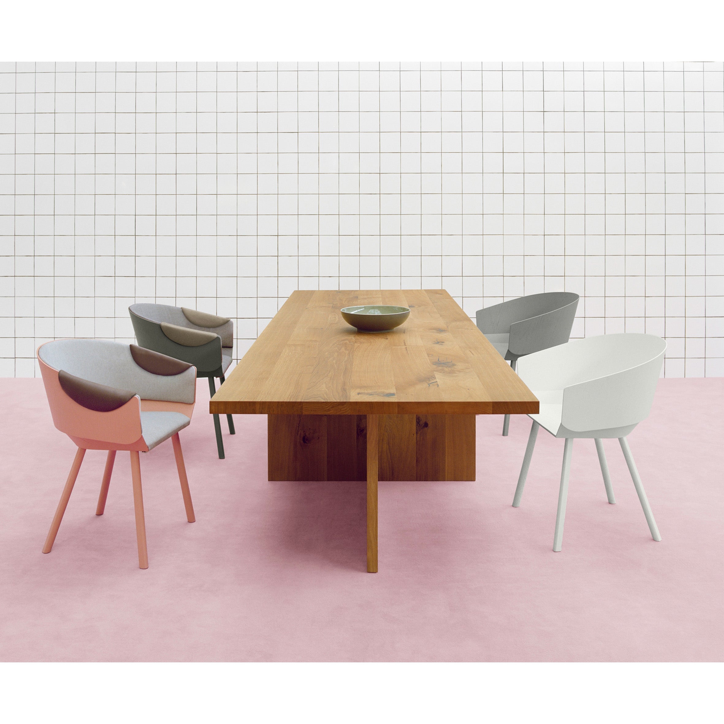 An E15 TA18 ZEHN table, made from European oak, is flanked by five modern chairs in pastel pink, gray, and white. A ceramic bowl graces its center. The backdrop includes a white grid-patterned wall and pale pink floor.