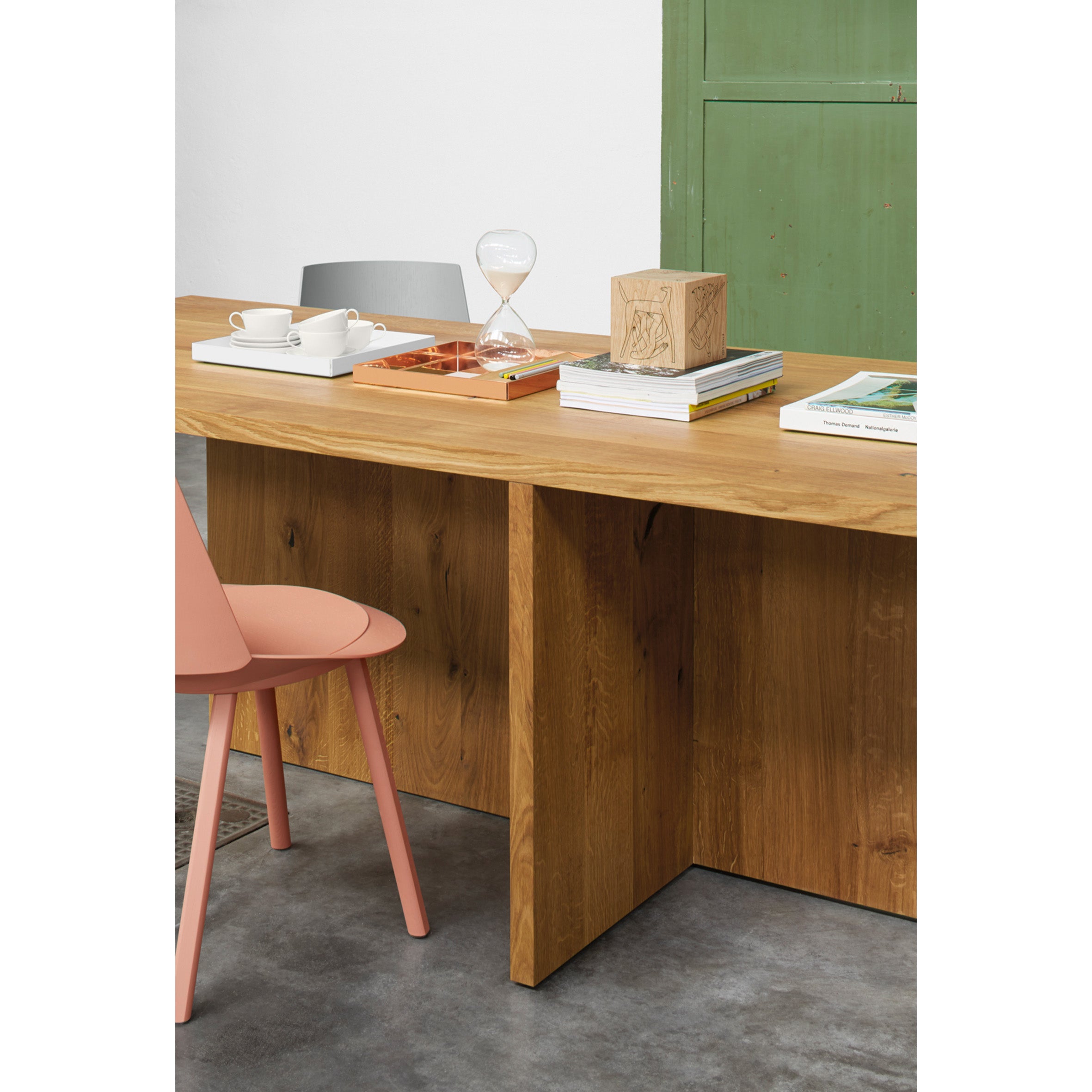 The TA18 ZEHN, a European oak table by E15, holds books, a sand timer, and a tray with a white cup and saucer. A pink chair complements the desk on the polished concrete floor, with a partially visible green cabinet in the background.