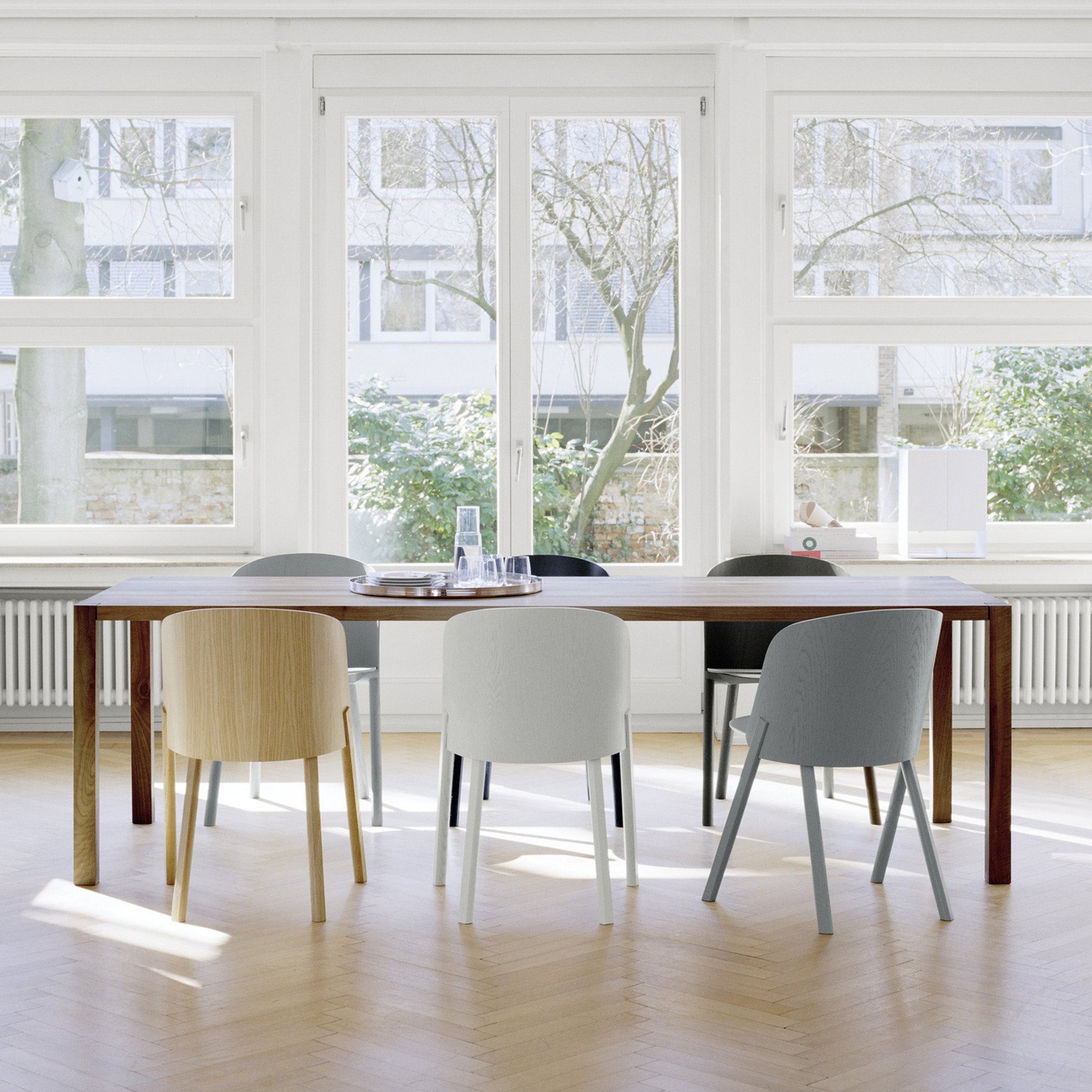 A modern dining space features the E15 TA19 SLOANE solid wood table, encircled by six minimalist light wood, white, and grey chairs. Large windows reveal trees and a building, with natural light highlighting the elegant design and herringbone flooring.