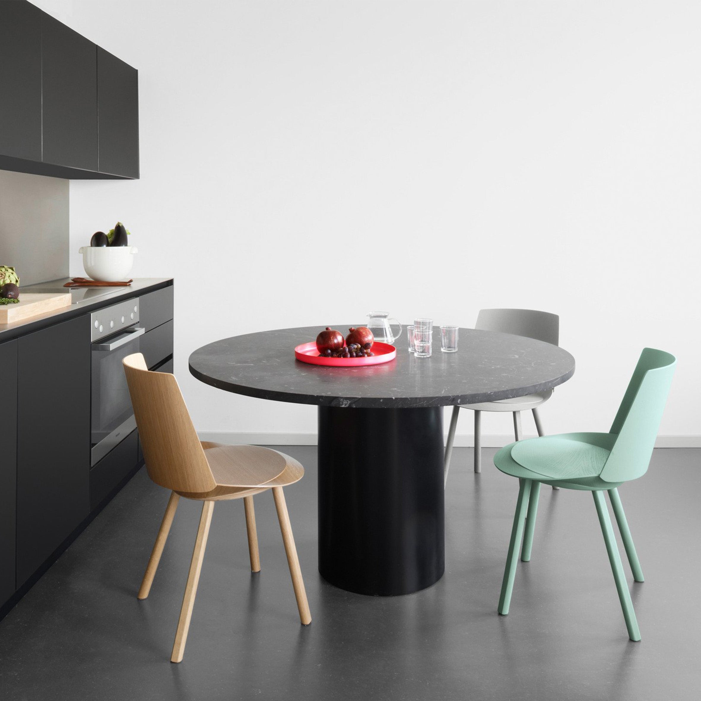 The modern kitchen showcases E15s sculptural black TA20 HIROKI round table, paired with three wood, gray, and mint green chairs. A red tray with apples and glasses decorates the table. The sleek black countertop featuring an oven enhances its versatile HIROKI charm.