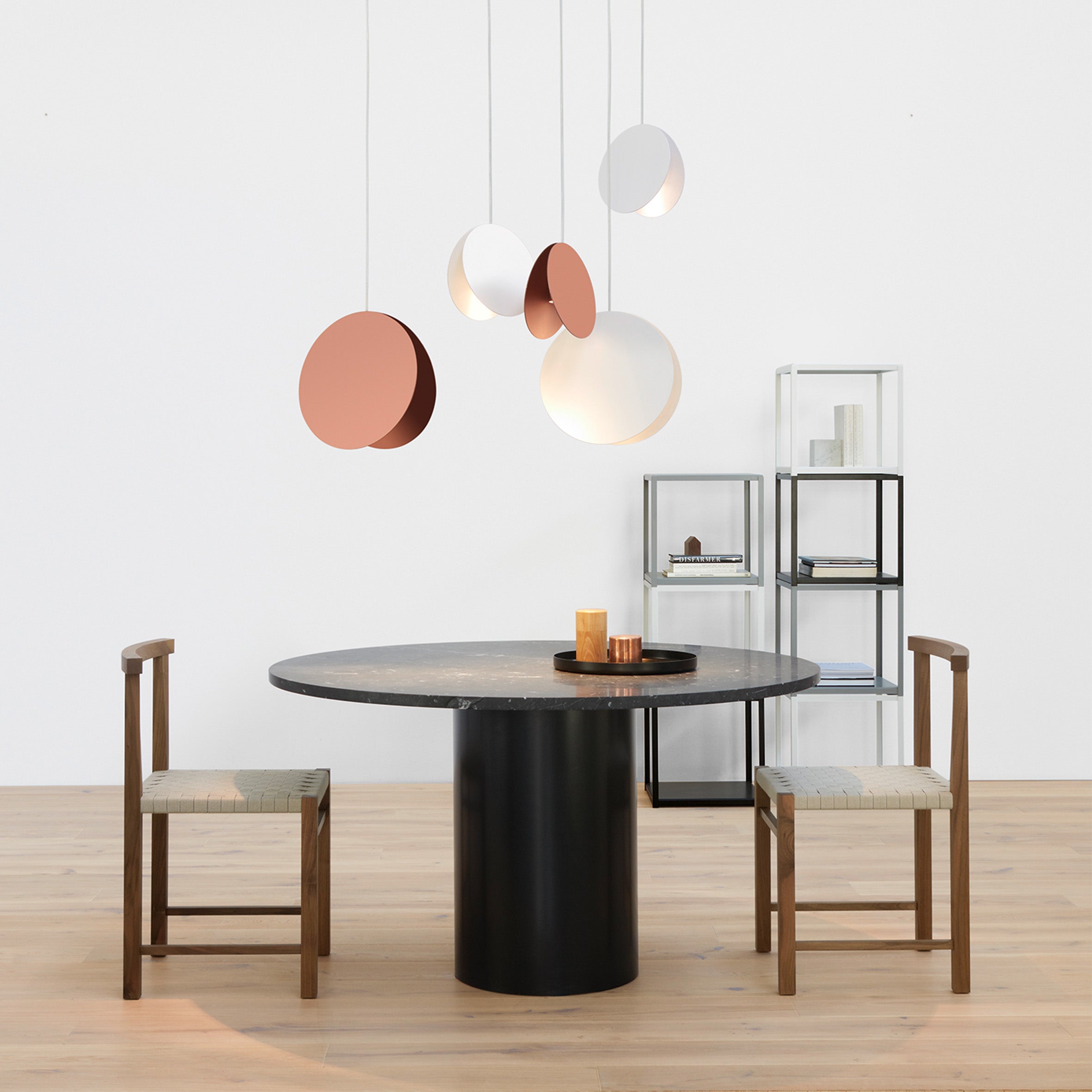 A minimalist dining setup showcases the sculptural TA20 HIROKI table and two wooden chairs. Overhead, modern circular pendant lights in brown and white provide a gentle glow. A shelving unit with decor complements the plain white wall and light wood floor, enhancing the serene ambiance. Brand: E15.