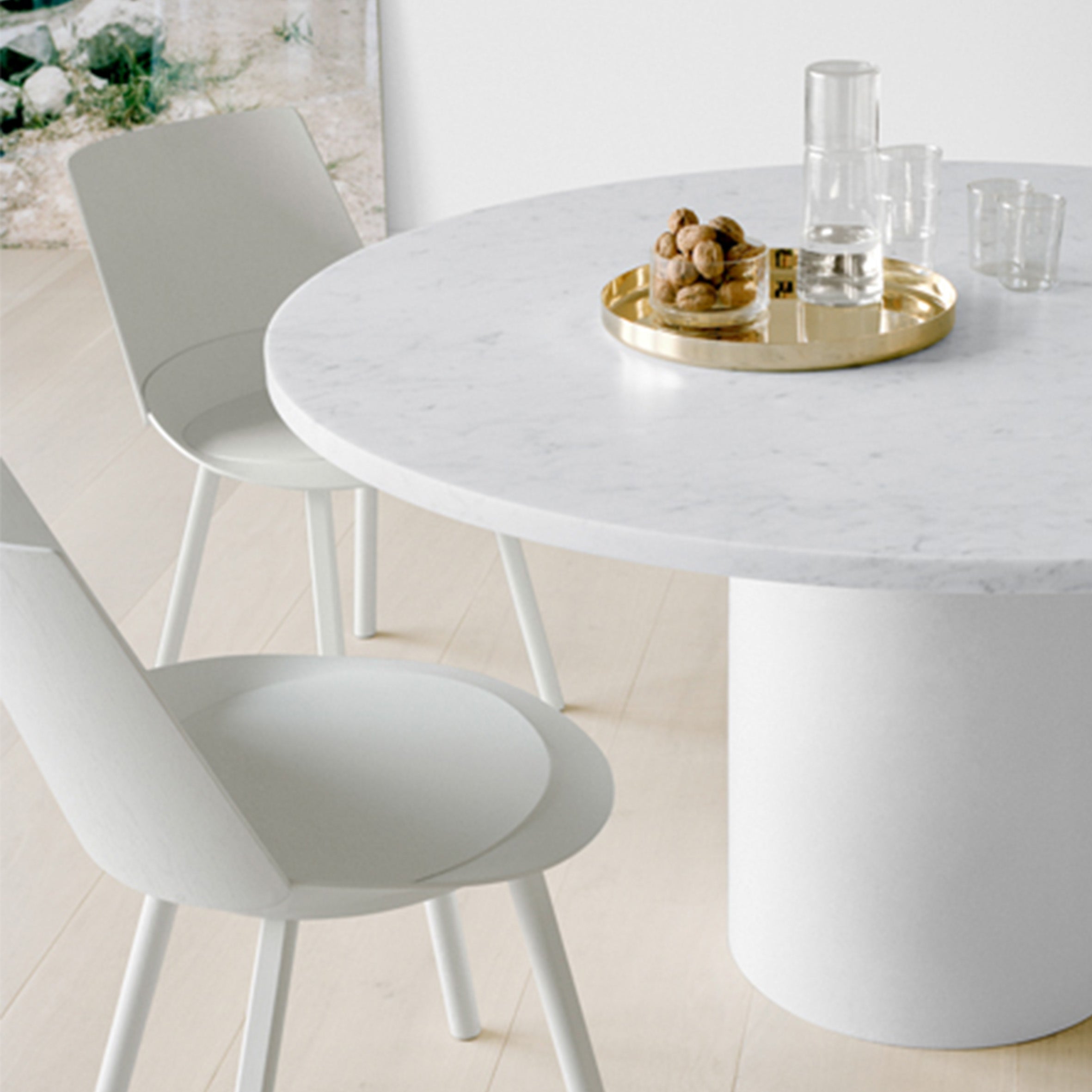 A minimalist dining setup features the sculptural TA20 HIROKI round table by E15, with a gold tray holding a glass carafe and glasses. Two white chairs surround it on light wooden flooring, partially revealing a green plant in the background.