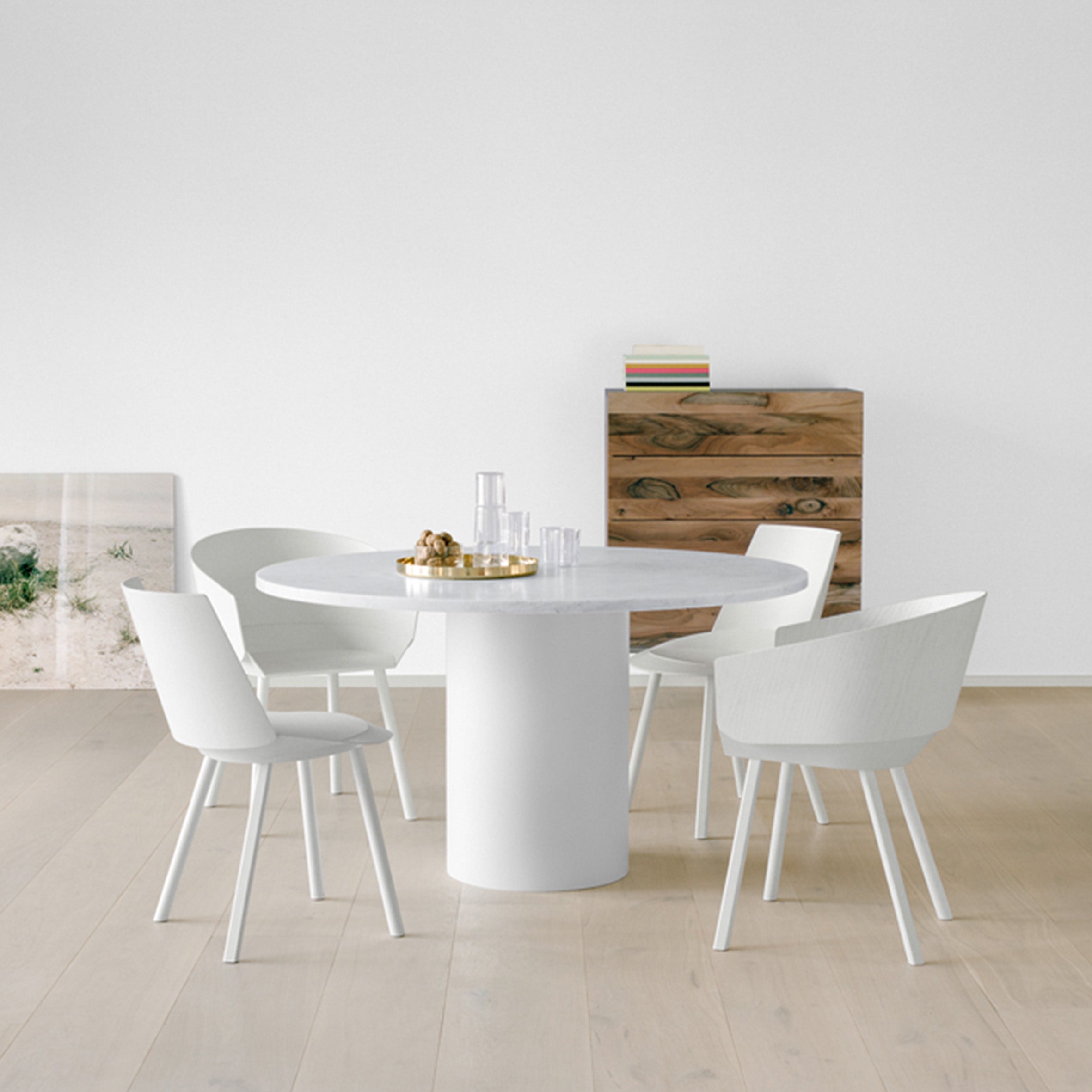 A modern dining area showcases E15s TA20 HIROKI round table with four white chairs. On the table sits a gold tray with bottles and glasses. A wooden chest and leaning artwork accent the light wooden floor, enhancing the versatile HIROKI ambiance.