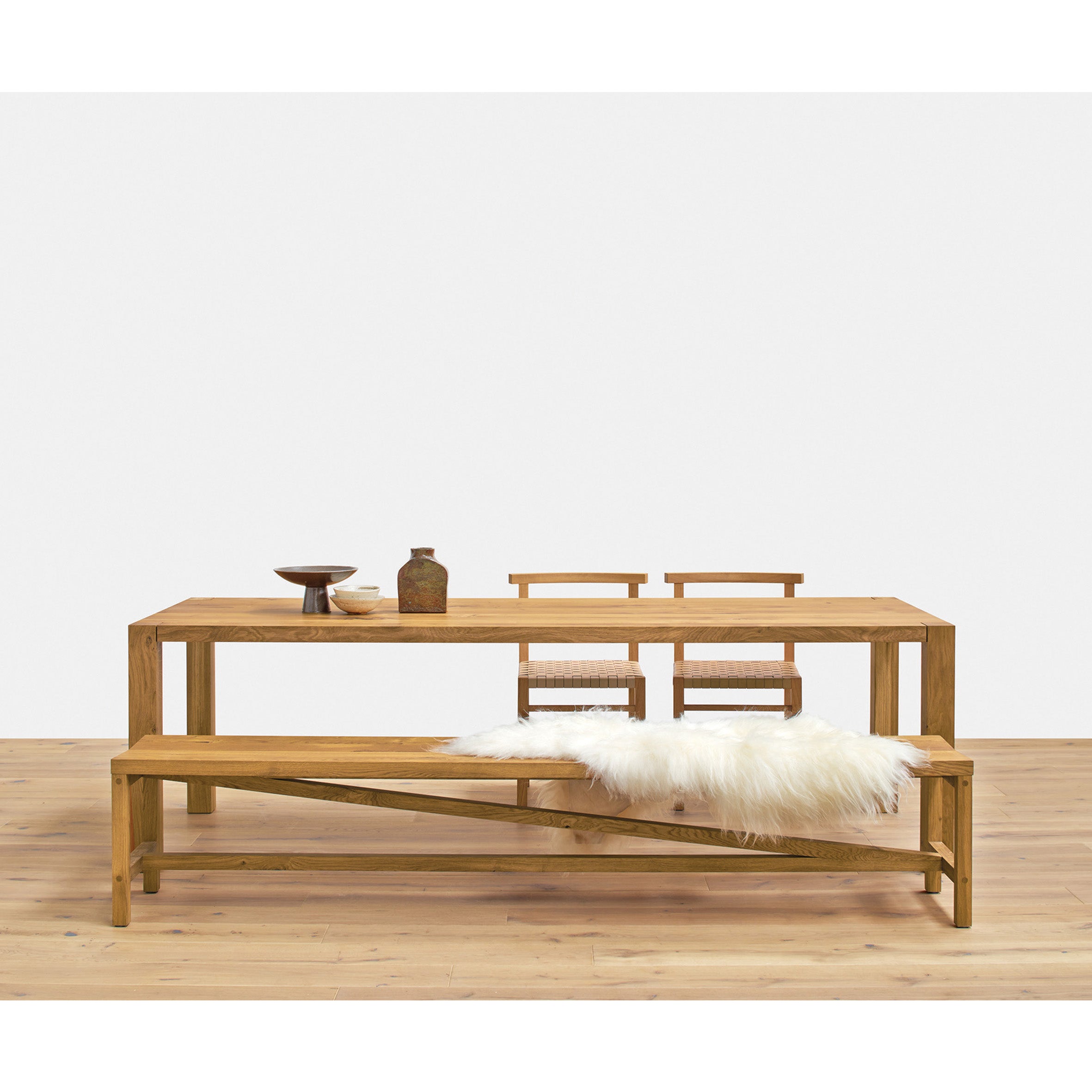 A minimalist dining setup features the E15 TA22 HOLBORN wooden dining table and chairs on light European oak flooring. A matching bench, adorned with a white fur throw, enhances the modern design, while a simple ceramic arrangement on the table adds to the serene ambiance.