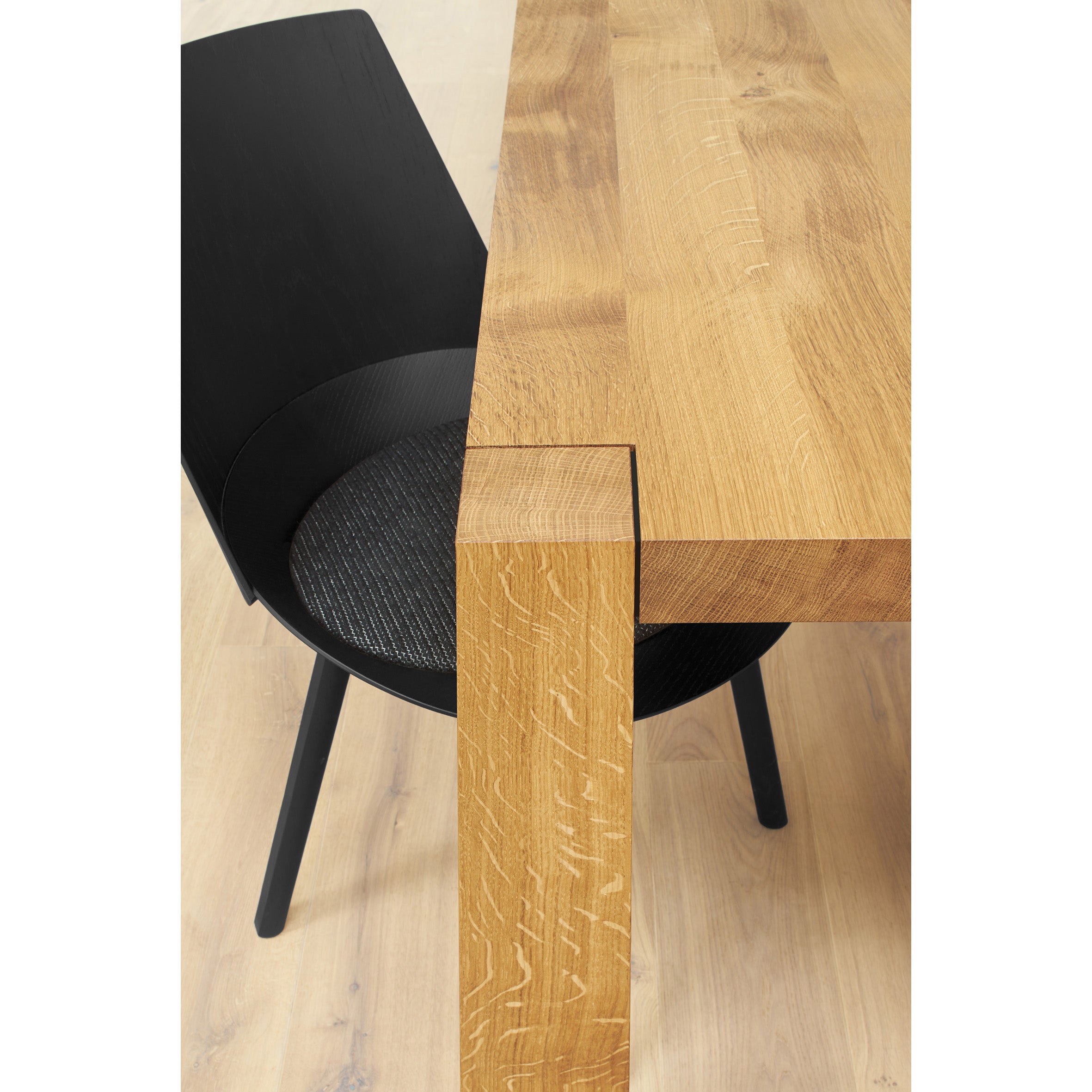 Close-up of a modern dining setup highlighting the E15 TA22 HOLBORN table made of thick, textured European oak with clean lines and a sleek black chair featuring a slightly curved backrest and seat cushion, placed on a light wood floor.