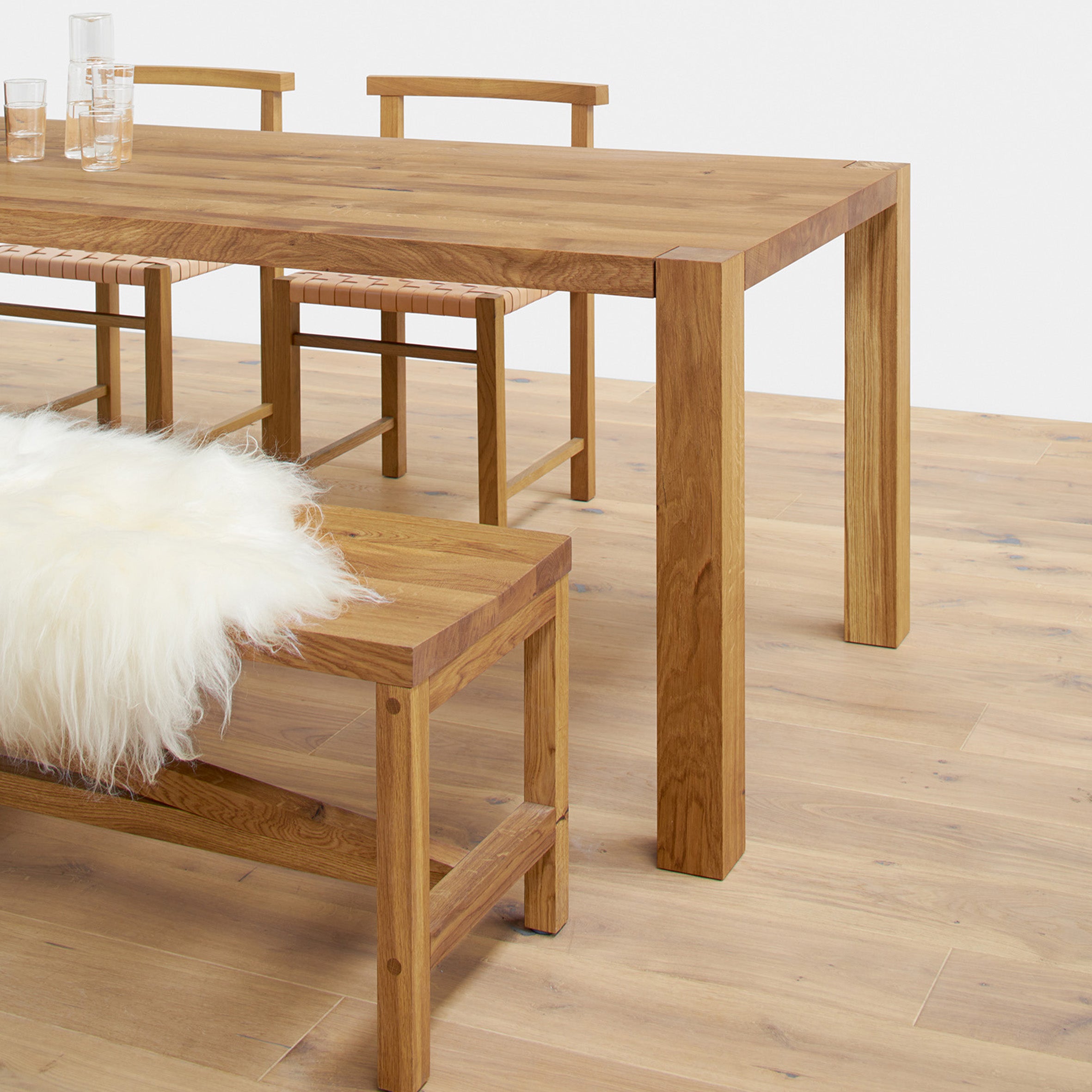 A minimalist dining area highlights the sleek E15 TA22 HOLBORN European oak dining table with matching chairs. A wooden bench adorned with a white fluffy throw complements the setup, while a light wood floor enhances the modern, cohesive, and natural design.