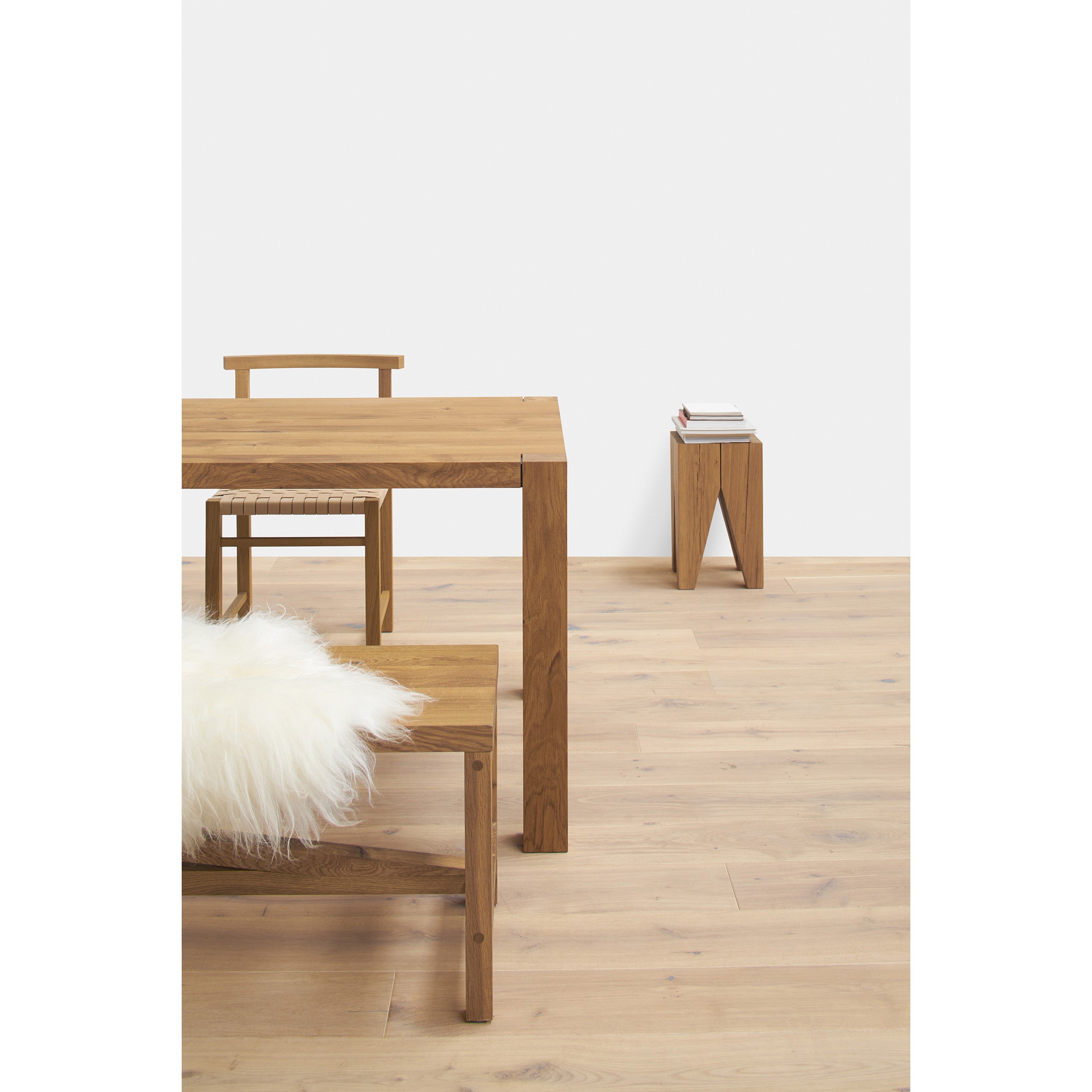 A minimalist room showcases the E15 TA22 HOLBORN modern European oak dining table, paired with a matching chair and bench featuring white fluffy cushions. A small side table holds a stack of books, while the light wood floor blends seamlessly with the plain white walls.