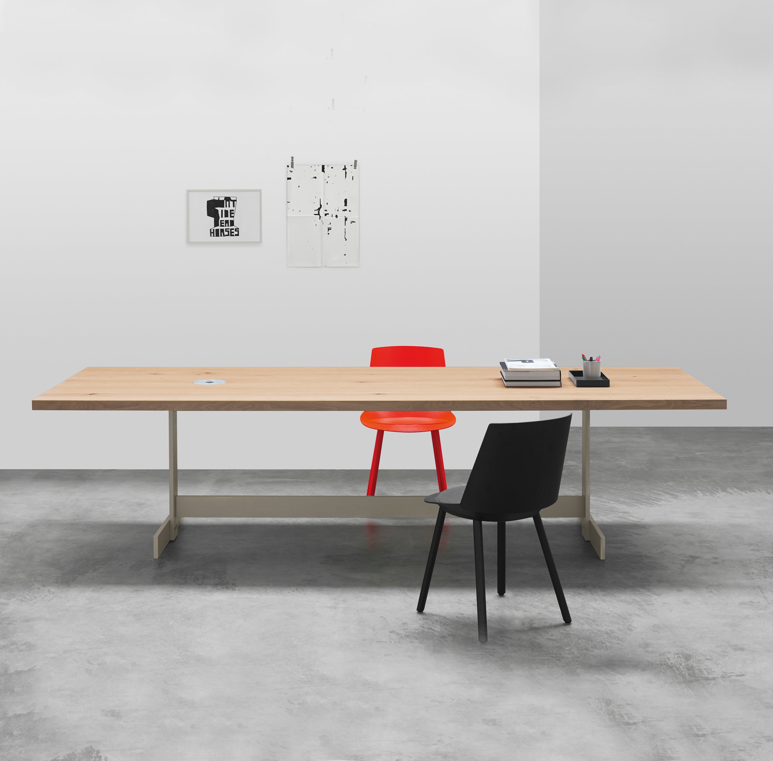 E15s TA23 KAZIMIR table made from solid European oak anchors the minimalist office with a red and black chair flanking it. The table showcases desktop essentials like books and a pencil holder, while two pieces of modern geometric art adorn the pristine white wall.