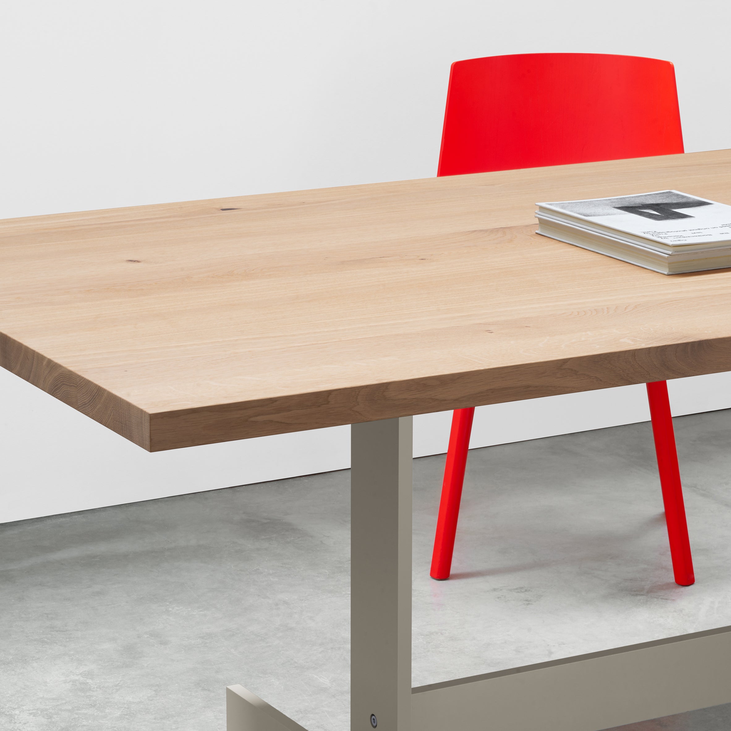 The E15 TA23 KAZIMIR, a modern wooden table crafted from solid European oak, stands in the foreground with a red chair tucked under it. Two magazines are stacked on top, highlighting its geometric design against a simple white wall and concrete floor.
