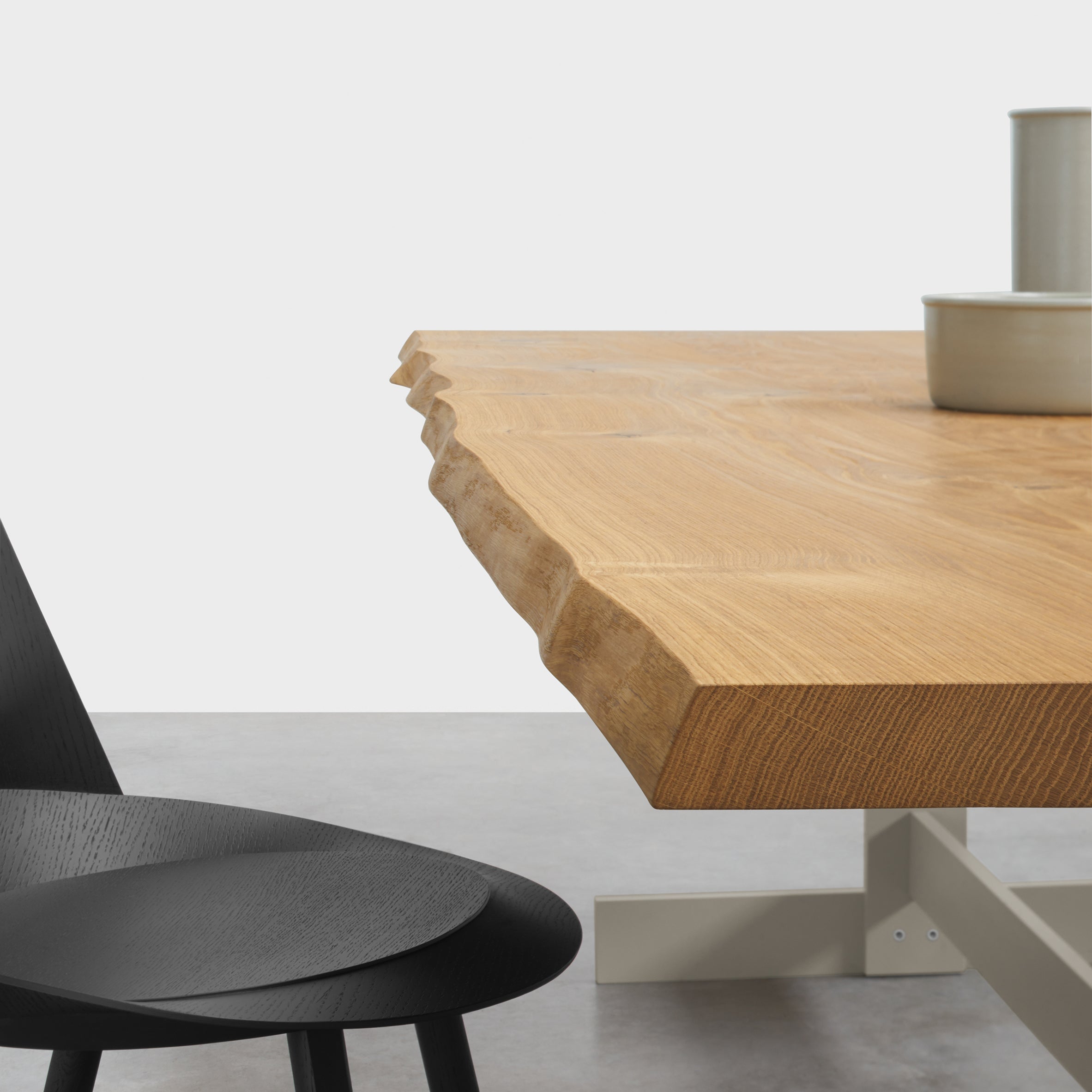 A close-up of the E15 TA23 KAZIMIR RAW table reveals a natural, jagged edge, crafted from solid European oak in light brown with visible grain patterns. To the side, a black chair with a curved backrest complements stacked white ceramic bowls on the tabletop.