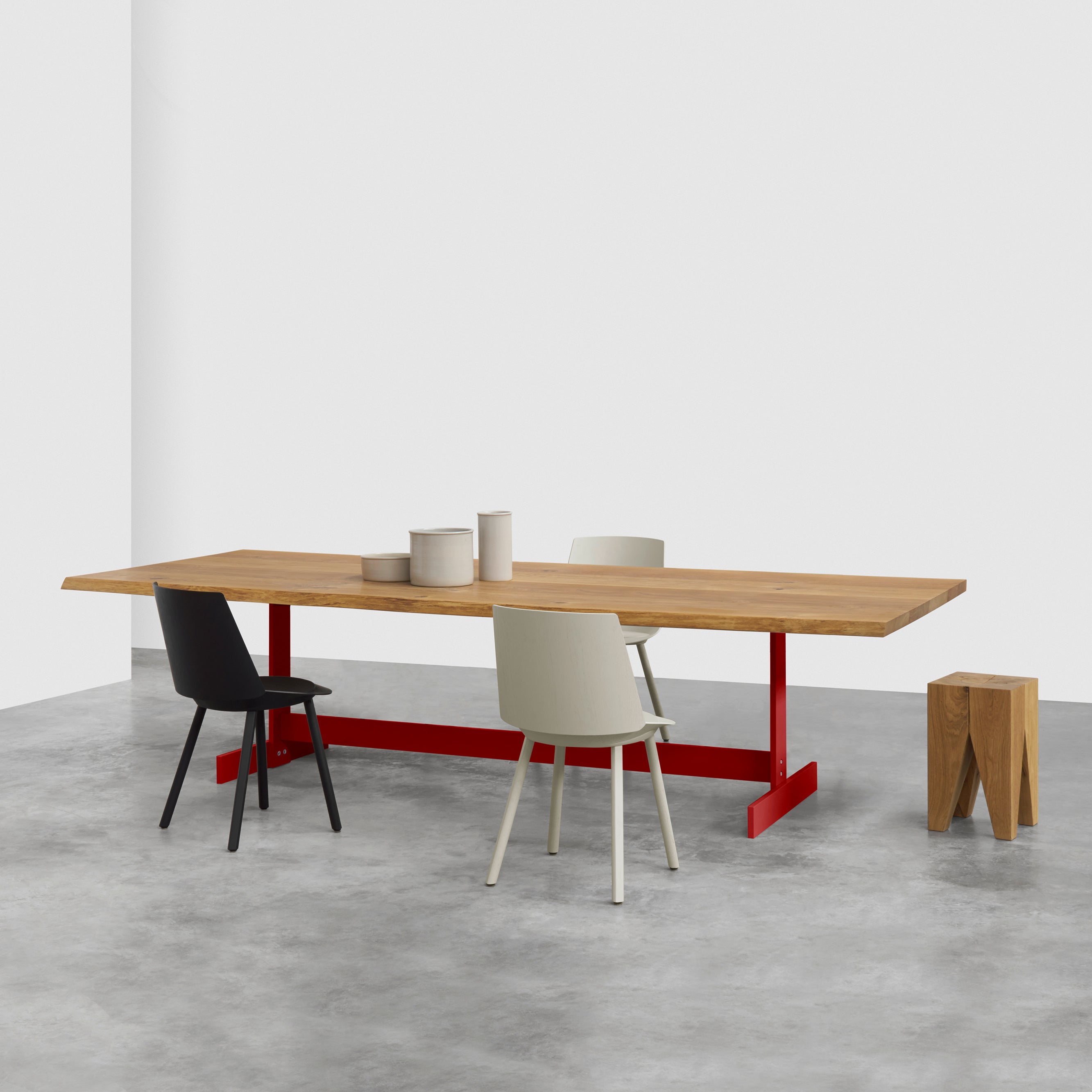 In a minimalist room, the TA23 KAZIMIR RAW table by E15, made from solid European oak with a red base, is paired with two black and white chairs and hosts three cylindrical vases. A small modern wooden stool rests nearby on the concrete floor.