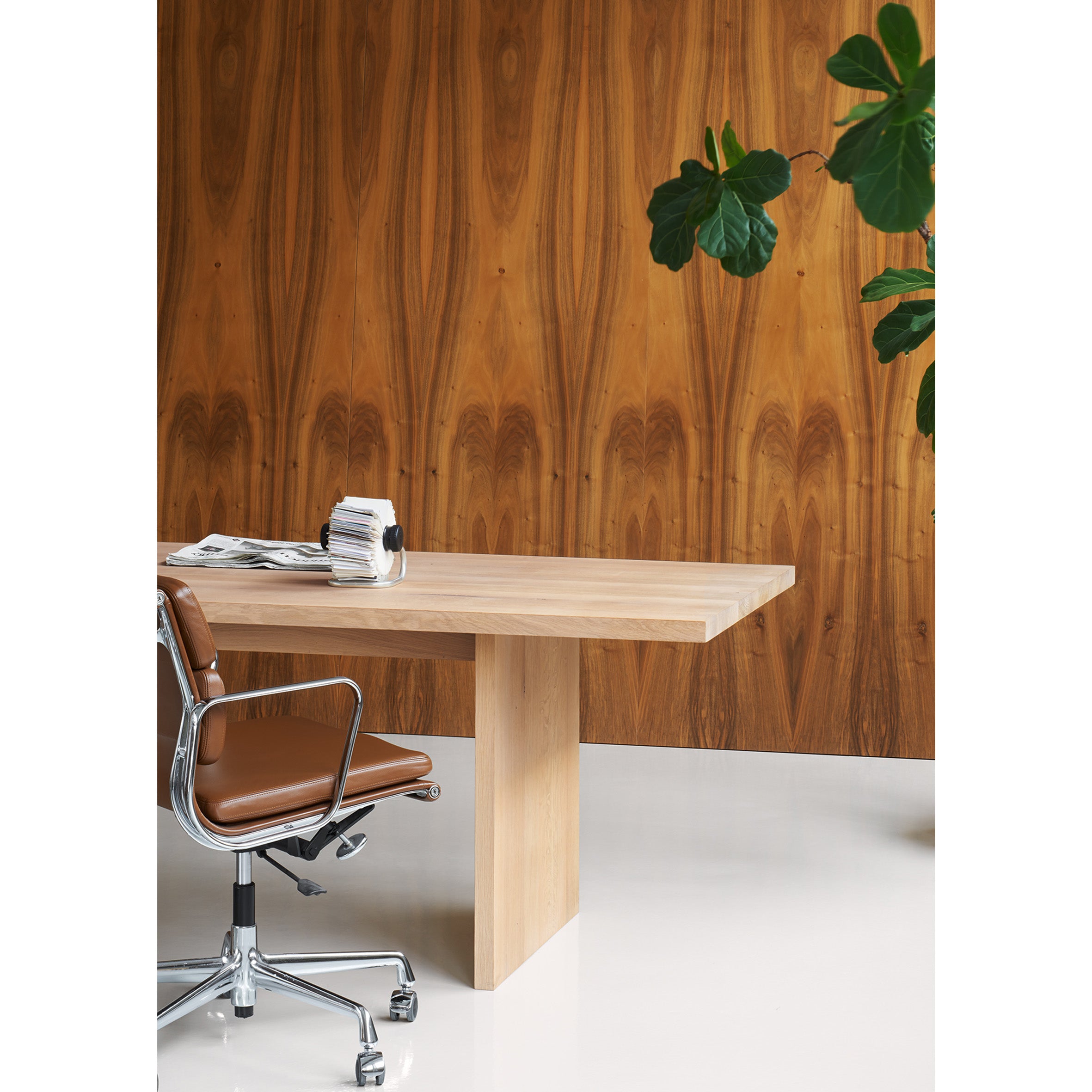 Minimalist office setup featuring the E15 TA24 ASHIDA solid wood desk and a brown leather swivel chair on a white floor. A sleek silver calendar rests on the desk. The backdrop showcases a Japanese carpentry-inspired wood-paneled wall and a large green potted plant.