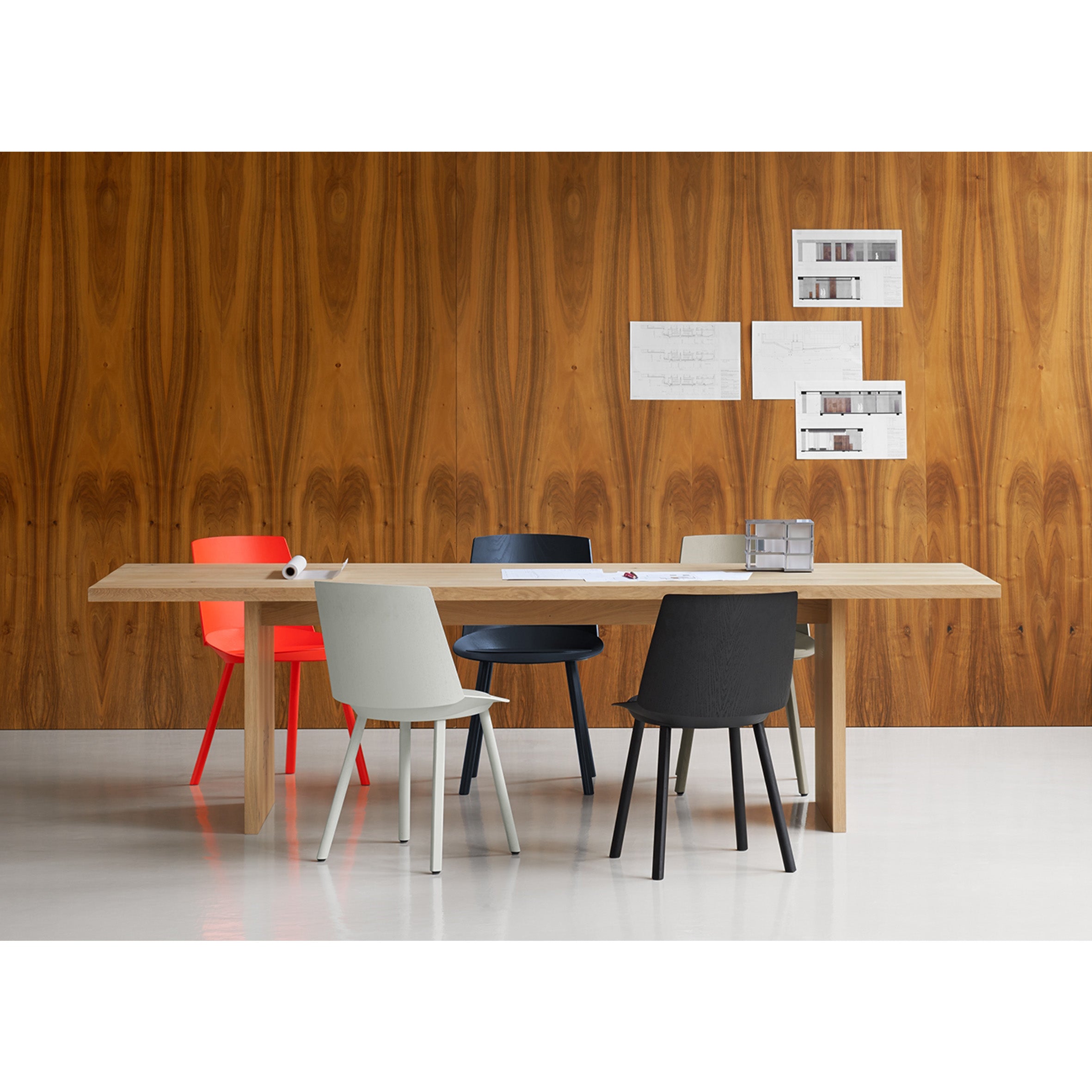The dining area features the E15 TA24 ASHIDA solid wood table, surrounded by chairs in red, beige, and black. The wall displays a natural grain pattern with three papers pinned to it. Crafted using Japanese techniques, the glossy white floor enhances the rooms elegance.