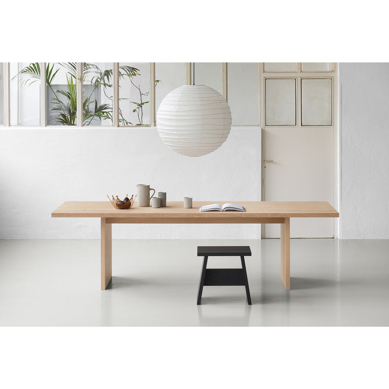 A minimalist room highlights the stunning TA24 ASHIDA solid wood table from E15, adorned with a bowl, jug, and open book. A black stool is placed below, while overhead a large white paper lantern brightens the space. Frosted windows and a white door frame tall plants, reflecting Japanese carpentry elegance.