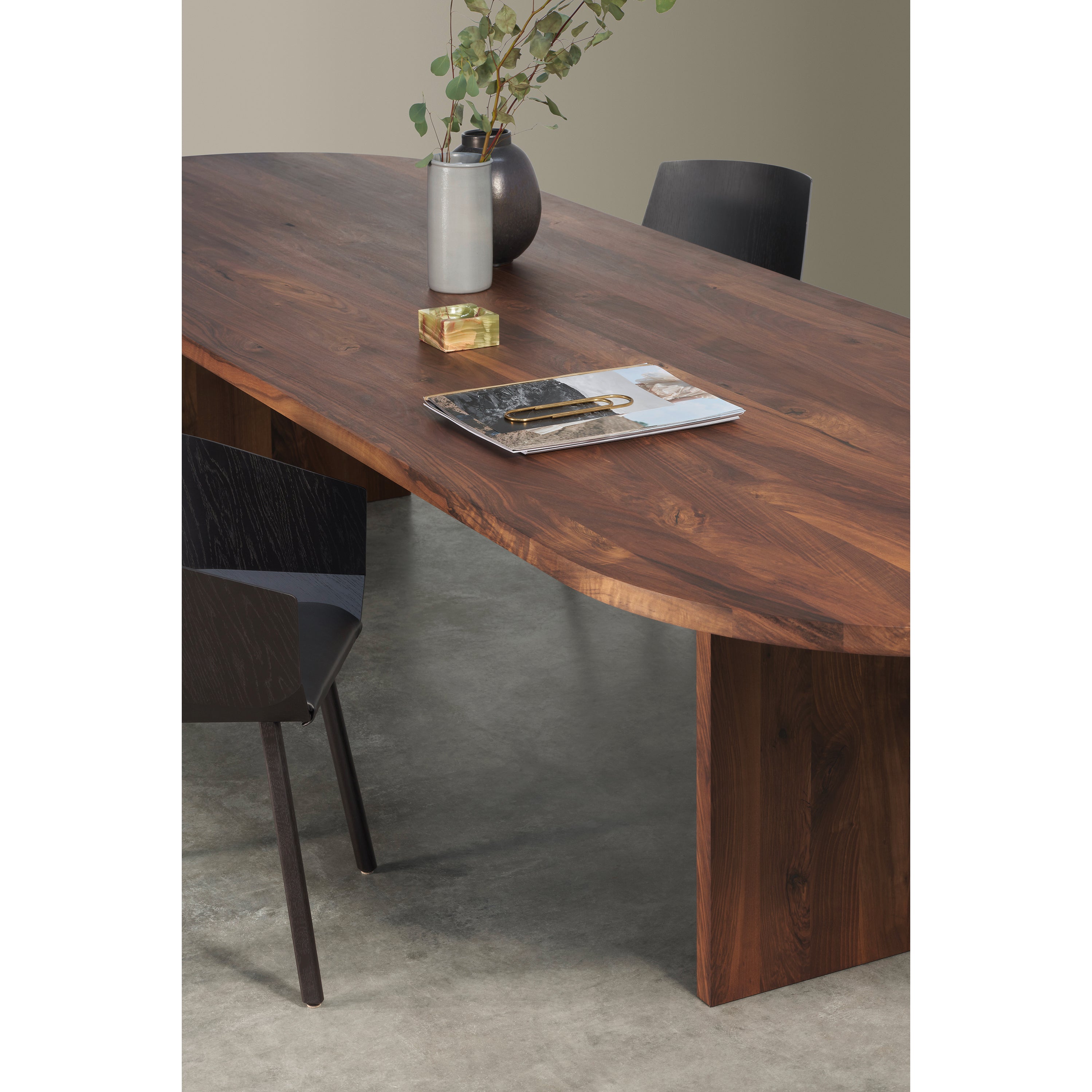 The TA24 ASHIDA Oval by E15 is a modern solid wood dining table made of European walnut with a smooth dark finish, surrounded by black chairs. It features two magazines, a glass vase with green branches, and a small gold box on top, complementing the rooms minimalist design and gray floor.