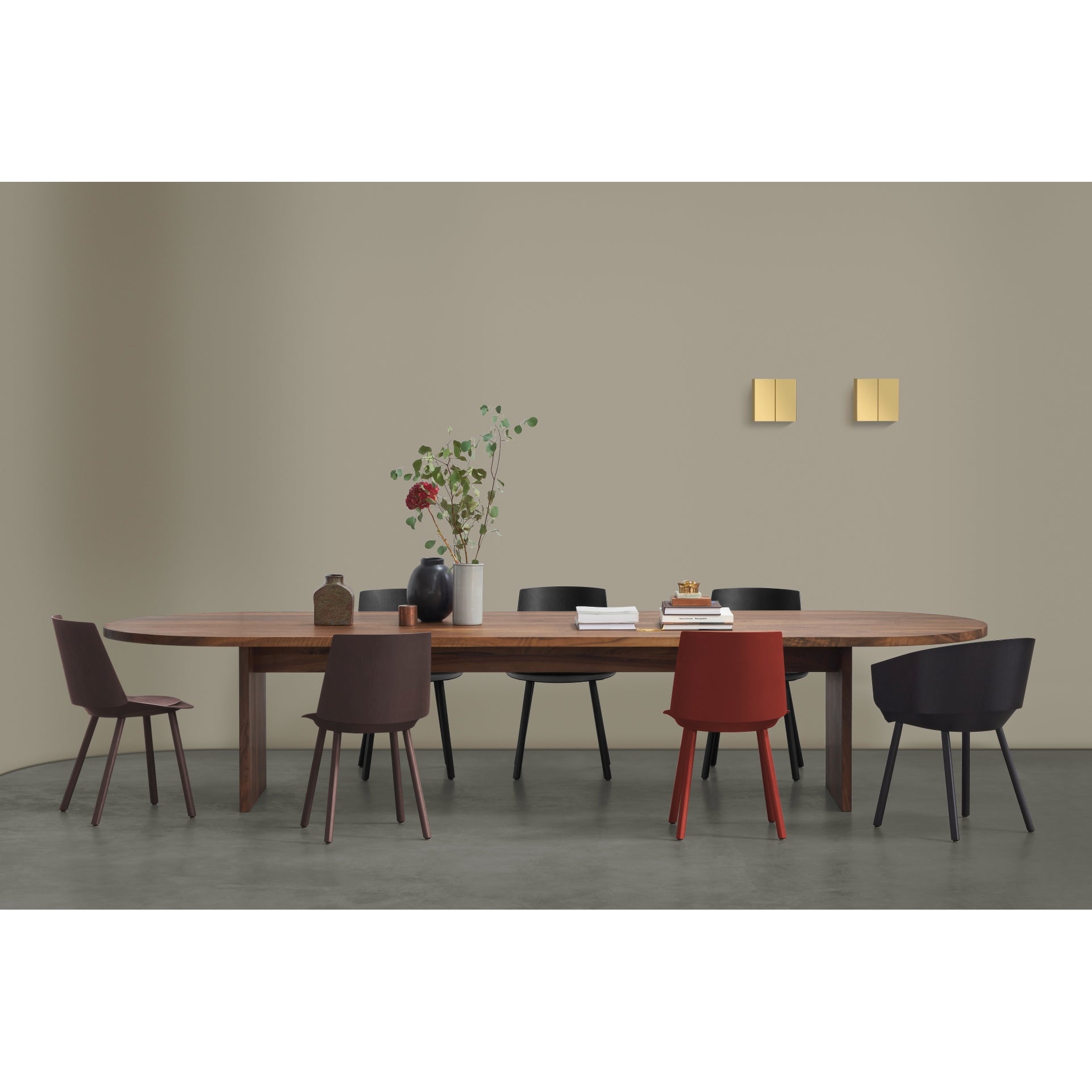 A contemporary dining room features E15s TA24 ASHIDA Oval solid wood table, encircled by black, brown, and red chairs. Two square wall lights illuminate the neutral walls, as floral vases enhance the tables elegance.