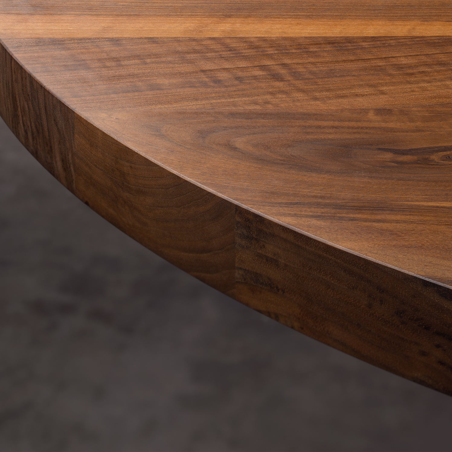 Close-up of the TA24 ASHIDA Oval table edge by E15, highlighting its smooth, polished surface with visible natural grain patterns. The blurred dark gray background accentuates the warm, rich tones of this stylish solid wood piece.