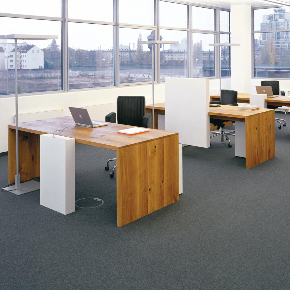 A modern office features wooden desks and black chairs on a gray carpet. One desk holds a laptop, papers, and an E15 BUSY wire manager. Large windows with powder-coated aluminum frames showcase cityscape views, buildings, and trees. The freestanding space is filled with bright natural lighting.