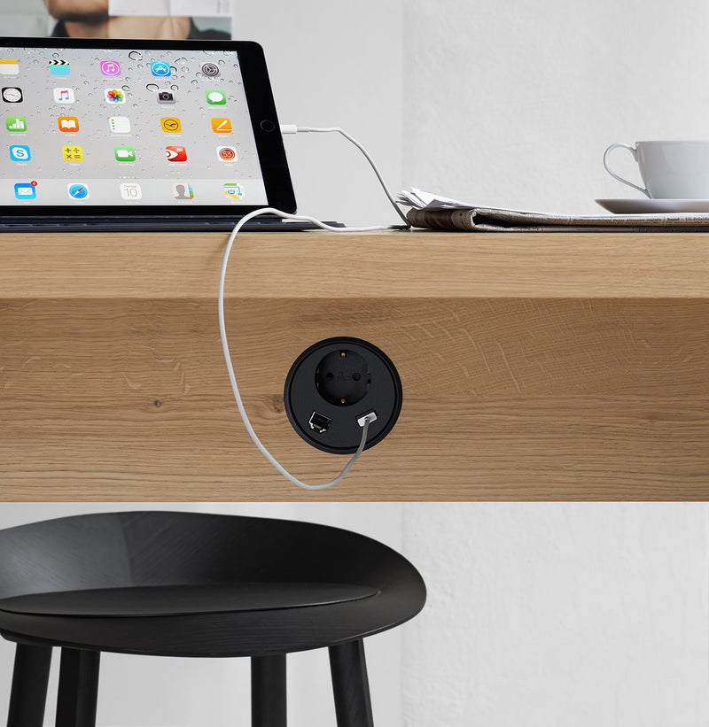 A tablet with a neatly plugged white charging cable demonstrates E15s excellent Wire Management on a wooden desk. Nearby, a newspaper and cup sit on the E15 table next to a sleek black chair.