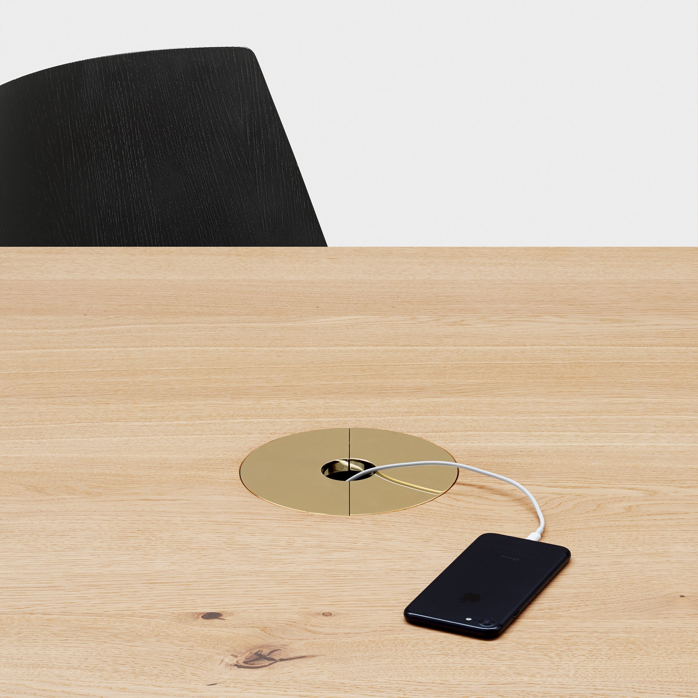 A black smartphone charges on an E15 wooden table, connected through a round, gold outlet. A black object peeks from the background. The minimalistic and modern setup highlights sleek wire management by E15 for a tidy look.