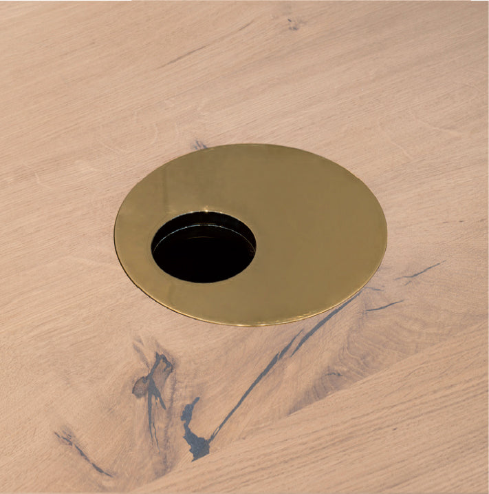 The E15 Wire Management solution features a polished brass grommet set in a light wood desk, blending visible grain patterns and darker knots with the sleek metallic finish for an elegant look.