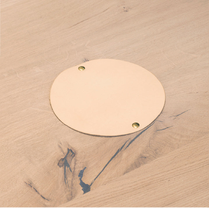 A light wooden surface features a round, beige coaster with brass studs. The E15 Wire Managements natural grain and subtle imperfections offer texture, blending form and function elegantly.