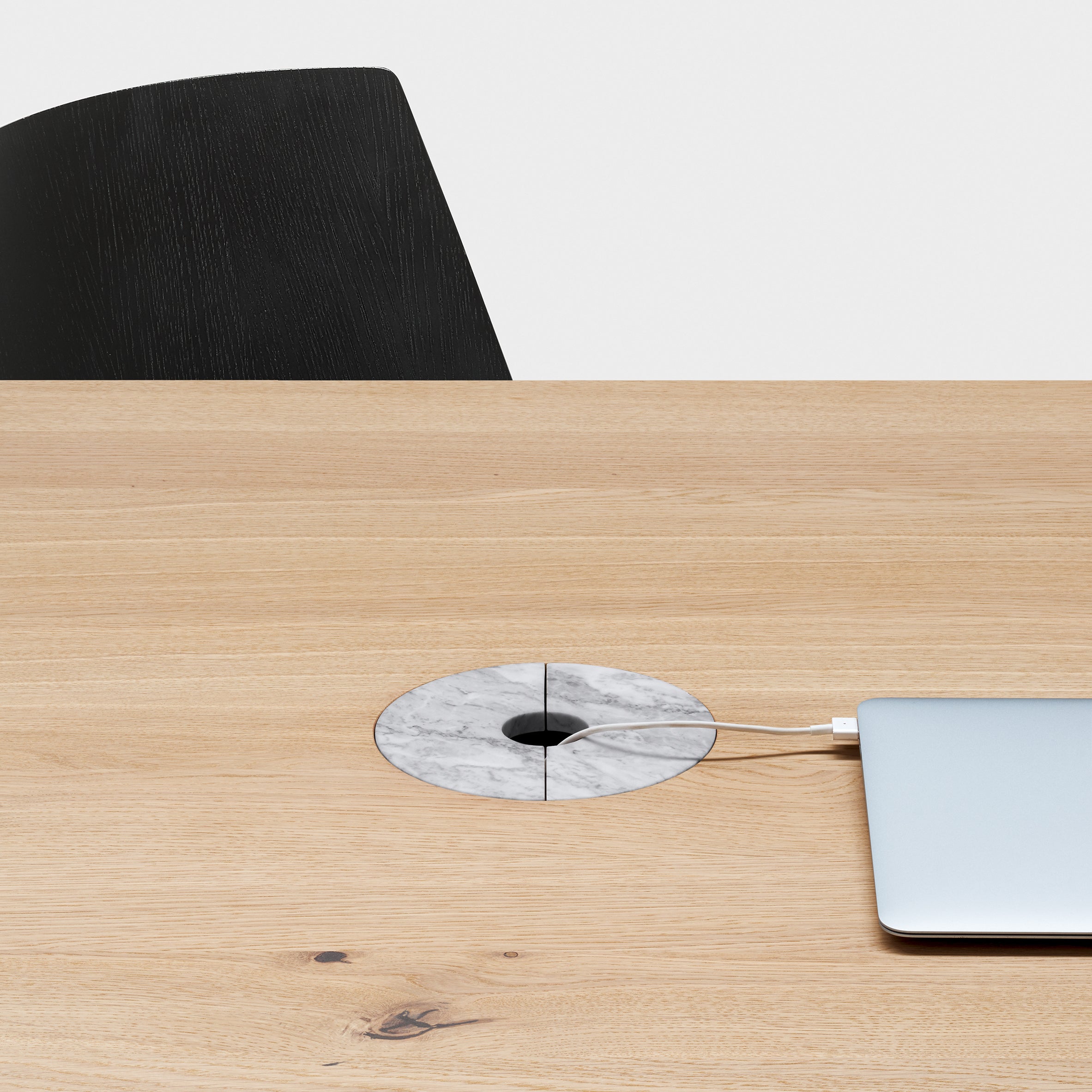 A minimalist workspace features an E15 table with Wire Management, round gray marble grommet for efficient cable organization. A silver laptop is partially visible on the right, and a black chair sits against a plain white wall in the background.