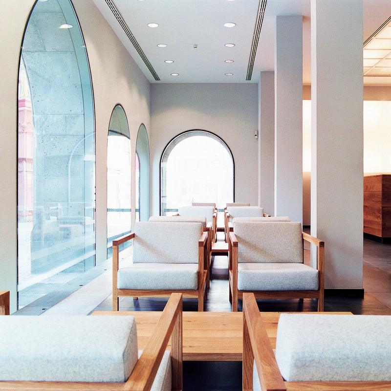 The modern cafe interior features large arched windows and solid wood accents. E15s EC01 BYRON wooden and white cushioned chairs, echoing classic smoking chairs, are neatly arranged in a row. Soft natural light fills the space, making it bright and inviting.