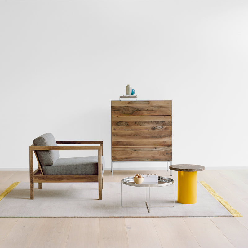 A minimalist room includes an E15 EC01 BYRON classic smoking chair with a gray cushion, a wooden dresser, gray rug, two small tables—one yellow cylindrical and one metallic with a fruit bowl—complementing the white walls and light wood floor.