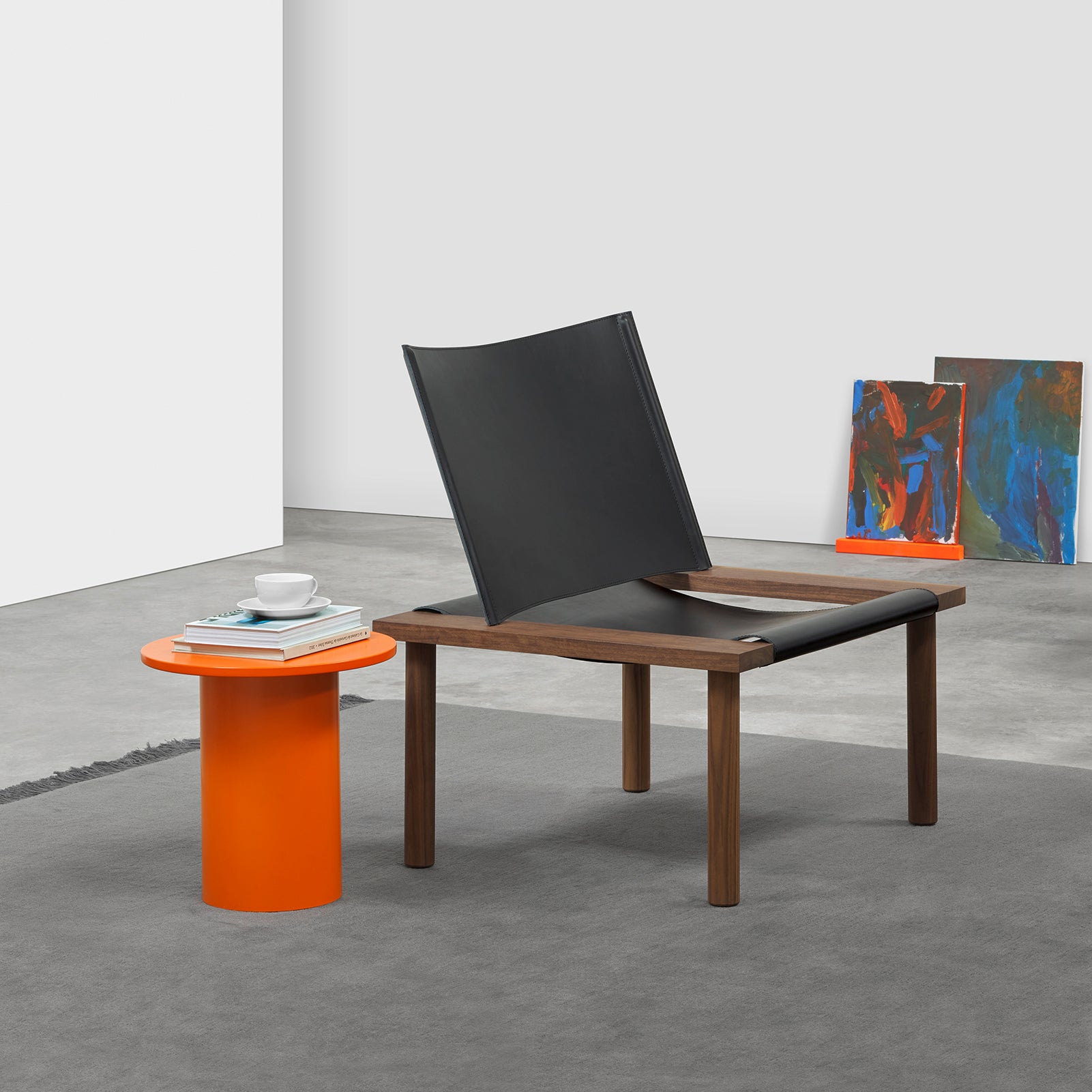 A modern room features an E15 EC06 ILMA Lounge Chair with a sleek black harness leather and solid wood frame. An orange cylindrical side table holds books and a cup. Gray carpet complements abstract paintings against the white wall, enhancing the elegance of natural materials.