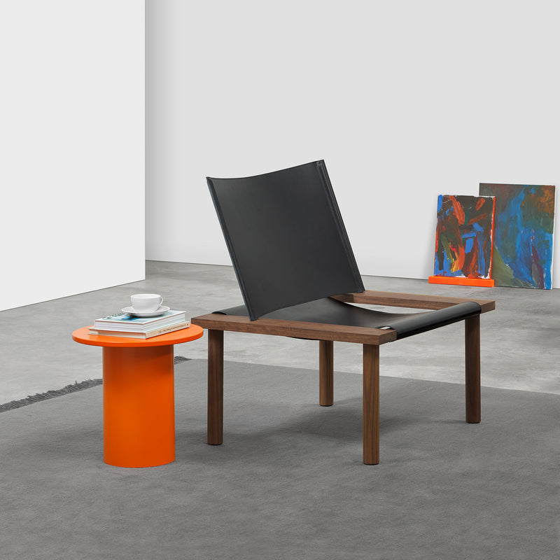 A modern room features an E15 EC06 ILMA Lounge Chair with a sleek black harness leather and solid wood frame. An orange cylindrical side table holds books and a cup. Gray carpet complements abstract paintings against the white wall, enhancing the elegance of natural materials.