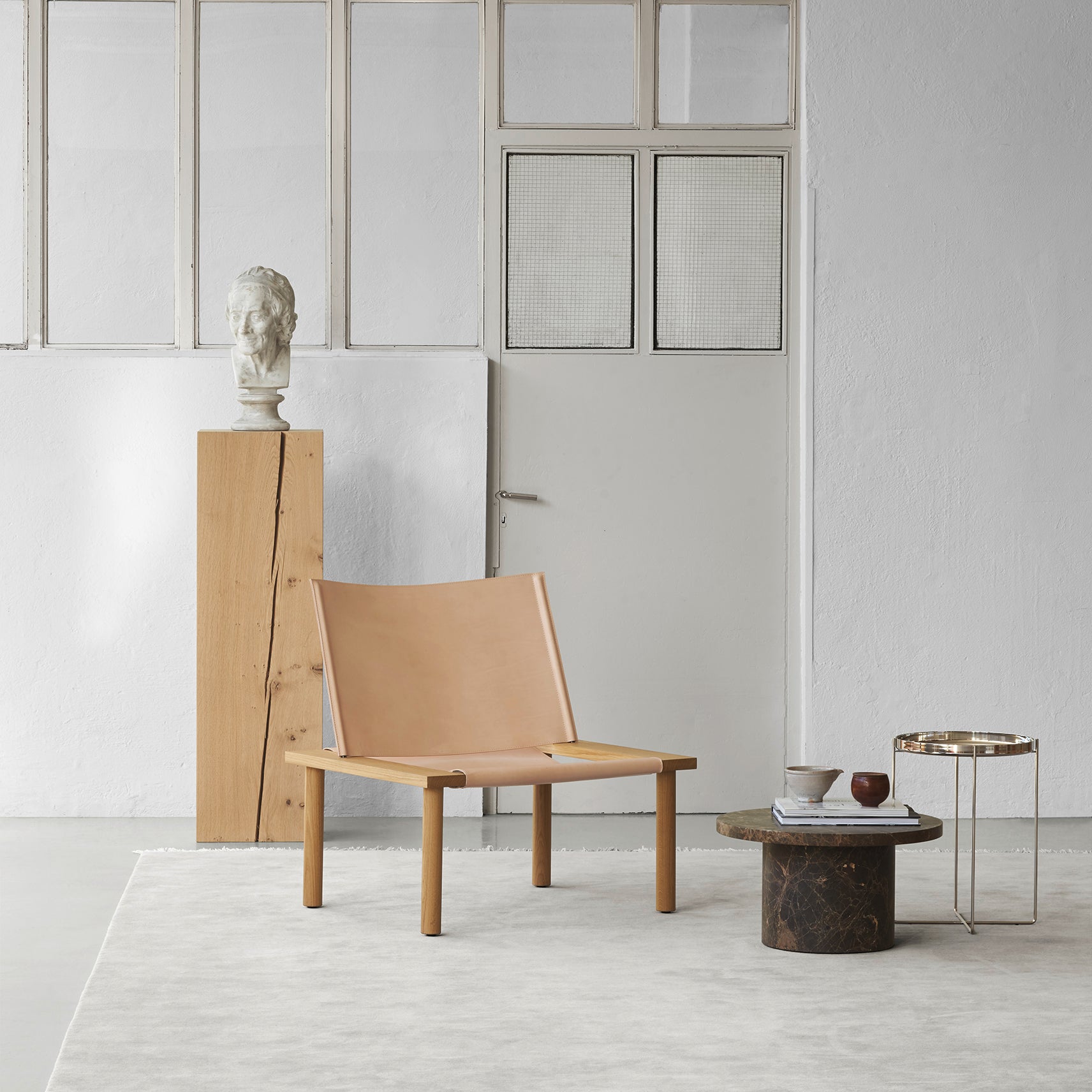 A minimalist room showcases the E15 EC06 ILMA Lounge Chair with a solid wood frame beside a small round table on a light rug. A tall pedestal displays a white bust, while glass-paneled doors in the background enhance the neutral palette, highlighting natural materials in its modern design.