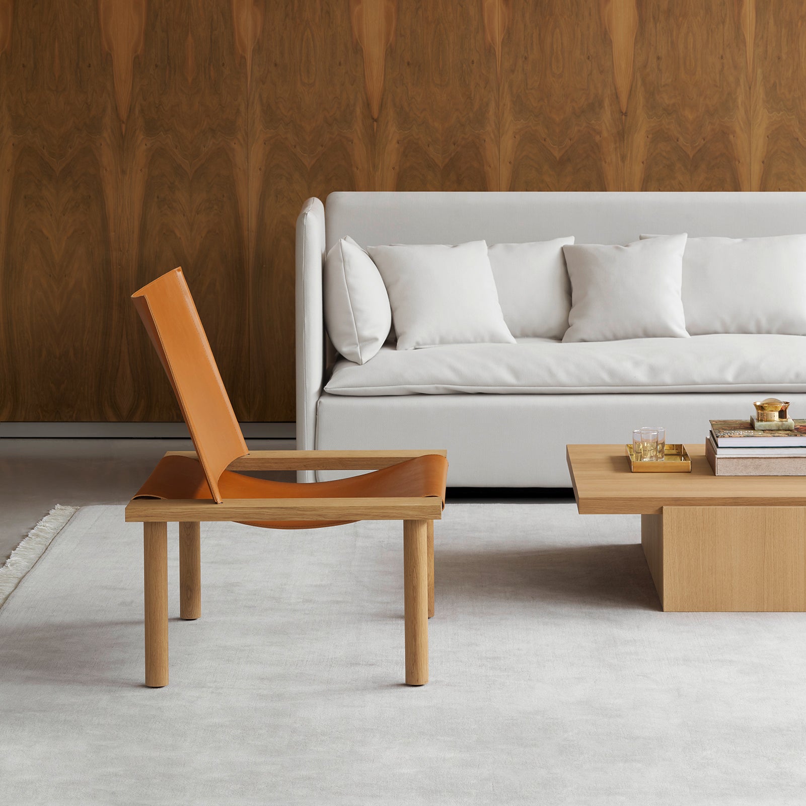 A modern living room features the EC06 ILMA Lounge Chair by E15 with a solid wood frame, a light gray sofa with white pillows, and a wooden coffee table displaying books and a glass tray. A neutral rug covers the floor, complemented by an elegant wood-patterned wall in the backdrop.