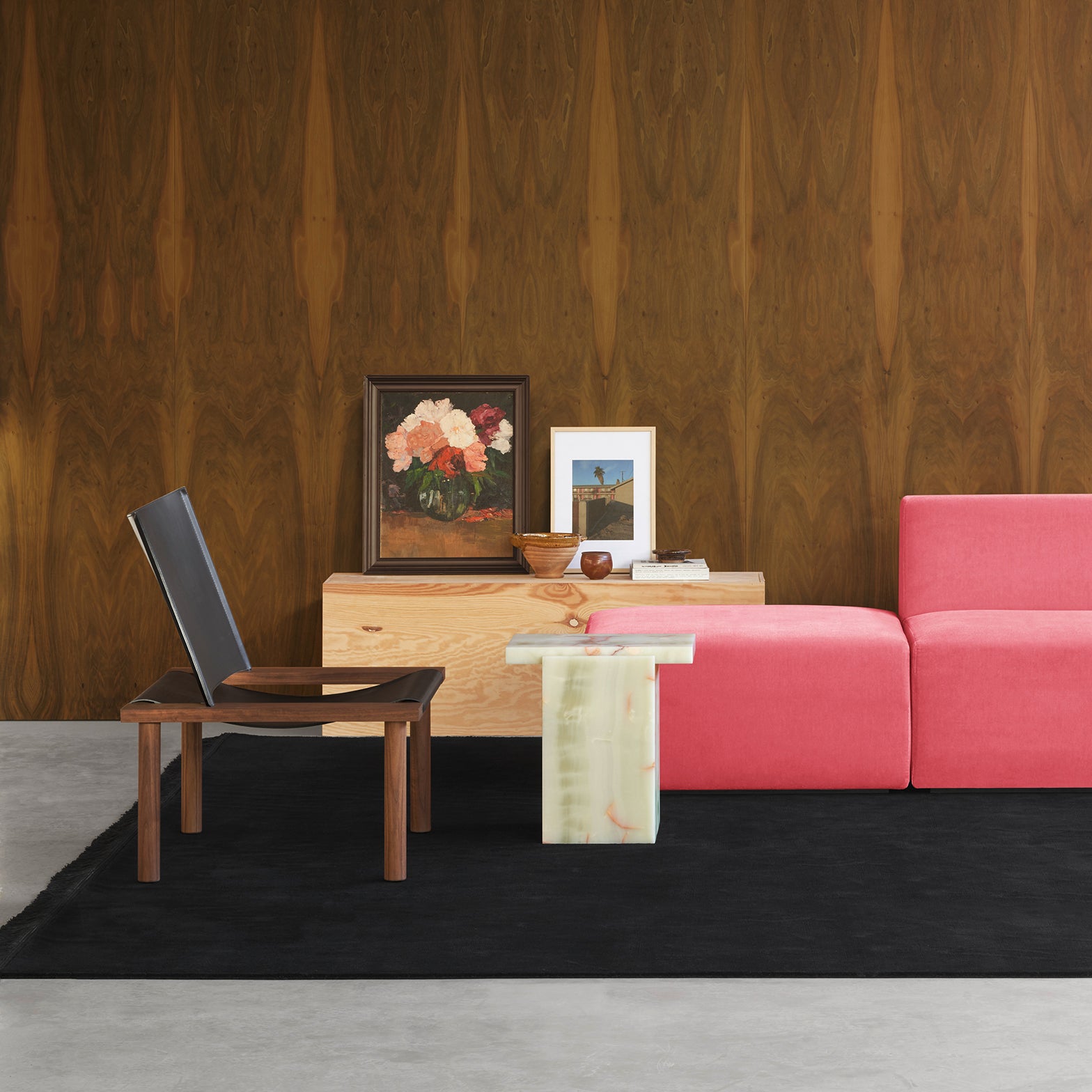 A modern living room showcases a pink sofa and the E15 EC06 ILMA Lounge Chair with solid wood frames. A marble-finished table displays art against a wooden-patterned wall, including a floral painting. A black rug on the gray floor complements the natural materials in the decor.