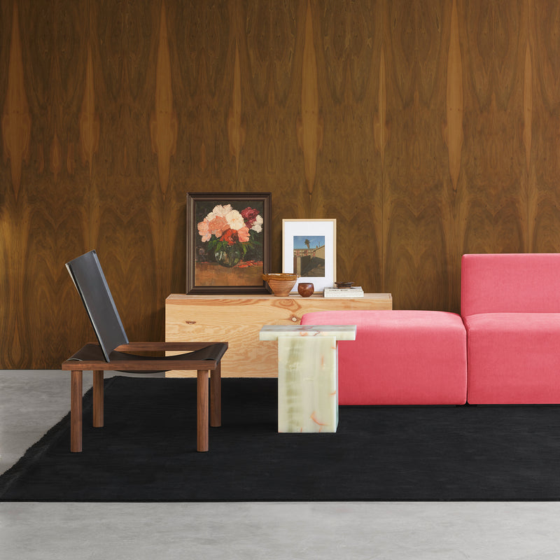 A modern living room showcases a pink sofa and the E15 EC06 ILMA Lounge Chair with solid wood frames. A marble-finished table displays art against a wooden-patterned wall, including a floral painting. A black rug on the gray floor complements the natural materials in the decor.