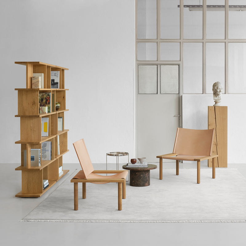 A minimalist room is highlighted by two EC06 ILMA Lounge Chairs from E15 on a light rug, alongside a wooden bookshelf filled with books and a small round side table. A pedestal with a bust enhances the décor, while large windows and a door accentuate the natural materials charm.