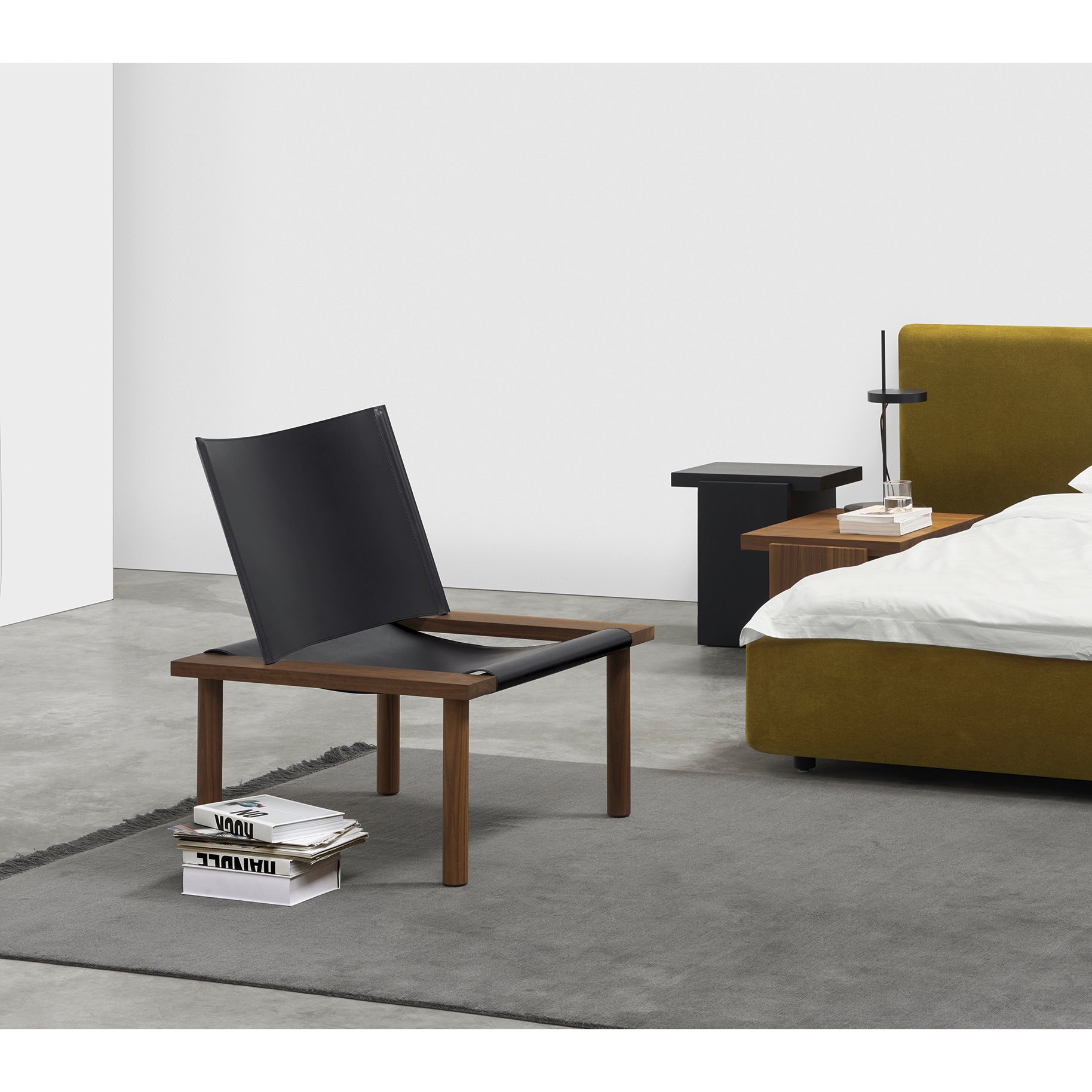 In a minimalist bedroom, E15s EC06 ILMA Lounge Chair with a black leather sling seat rests on a gray rug. A low bedside table holds a lamp and books beside a bed with an olive green upholstered headboard, all against concrete floors and white walls, emphasizing natural materials.