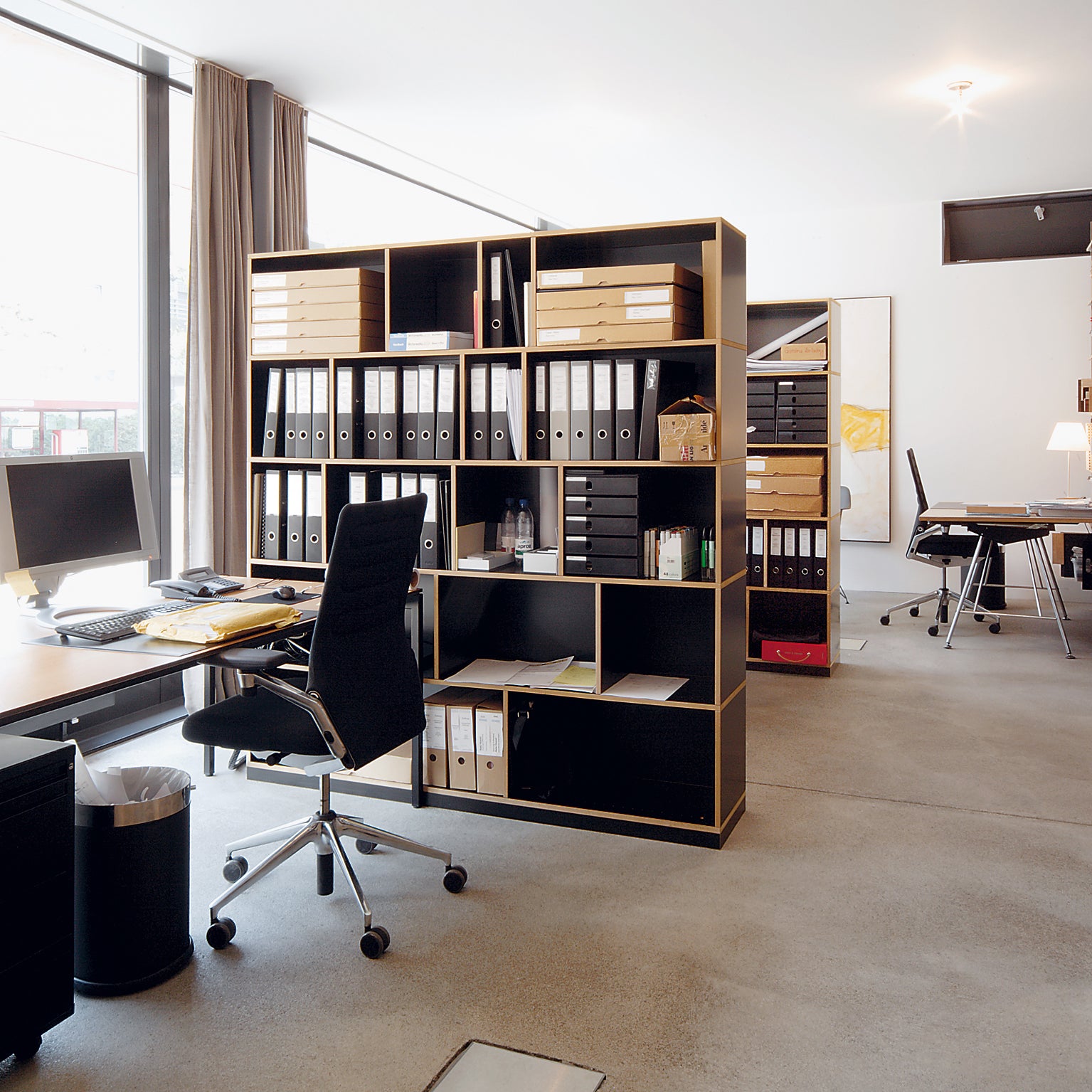 A modern office with large windows and sleek black chairs features a Moormann Egal shelf system dividing the space. The polished concrete floor enhances the minimalistic decor, flooded with natural light, while customizable shelving neatly organizes binders and boxes.