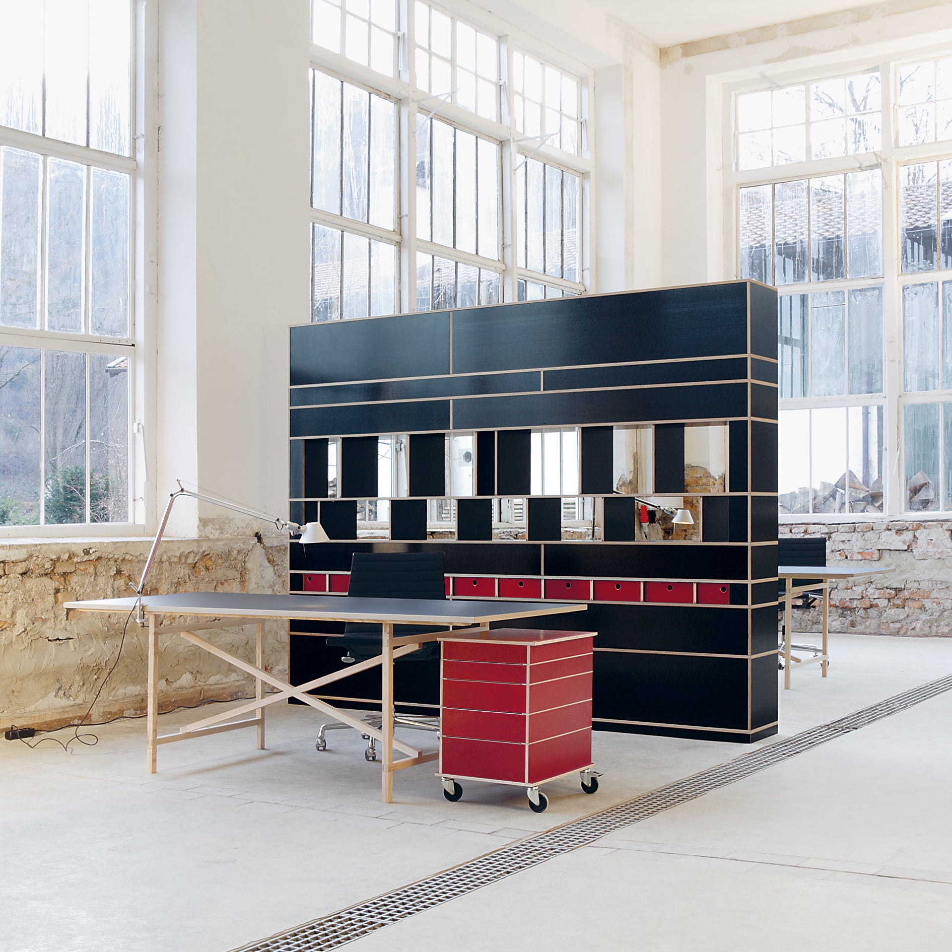 In a modern studio with large windows, a minimalist workspace is highlighted. A Moormann Egal black partition with customizable shelving and small red drawers is in the background, while a wooden desk with a task lamp and red wheeled drawer unit stands prominently in the foreground.