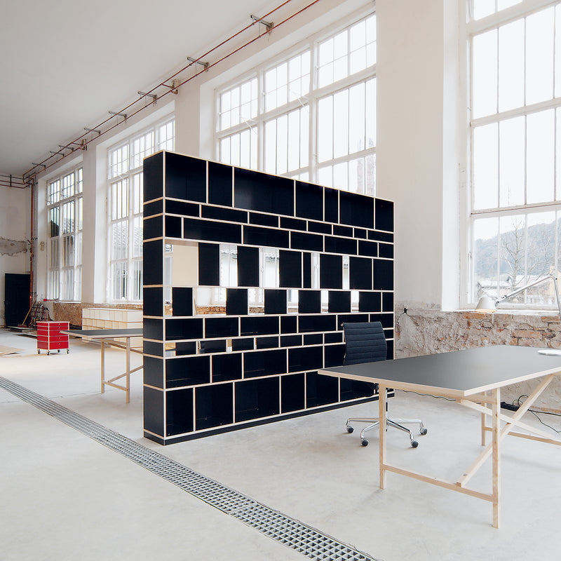 The spacious modern office boasts large windows, customizable geometric shelving, and wooden desks with black surfaces by Moormanns Egal collection. An office chair enhances the chic ambiance, exposed brick walls add an industrial touch, and natural light bathes the well-lit room.