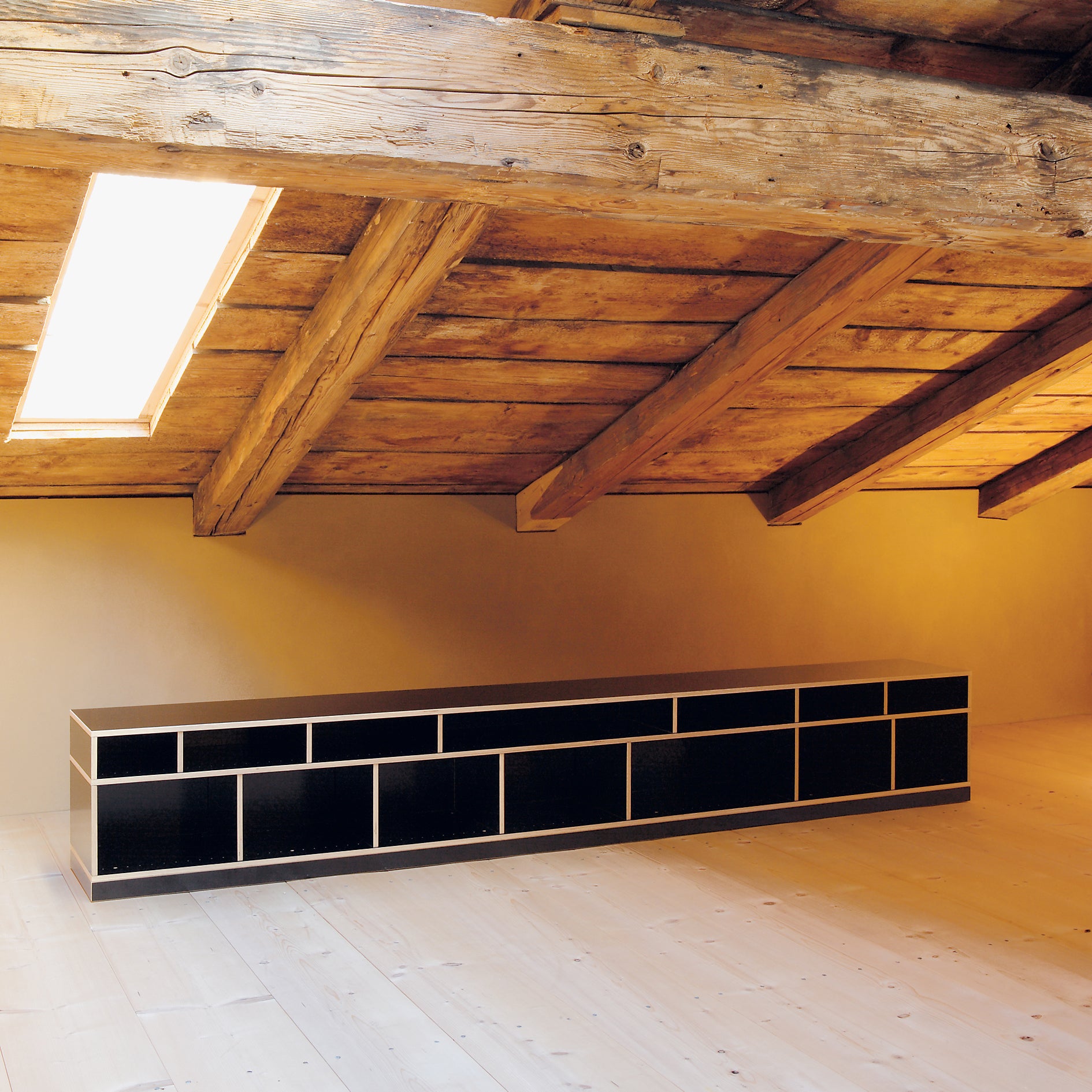 A minimalist room with a slanted wooden ceiling and plank floor is illuminated by sunlight from a skylight. Along the beige wall, Moormanns sleek Egal black cabinet with customizable shelving contrasts beautifully with the warm wood tones.