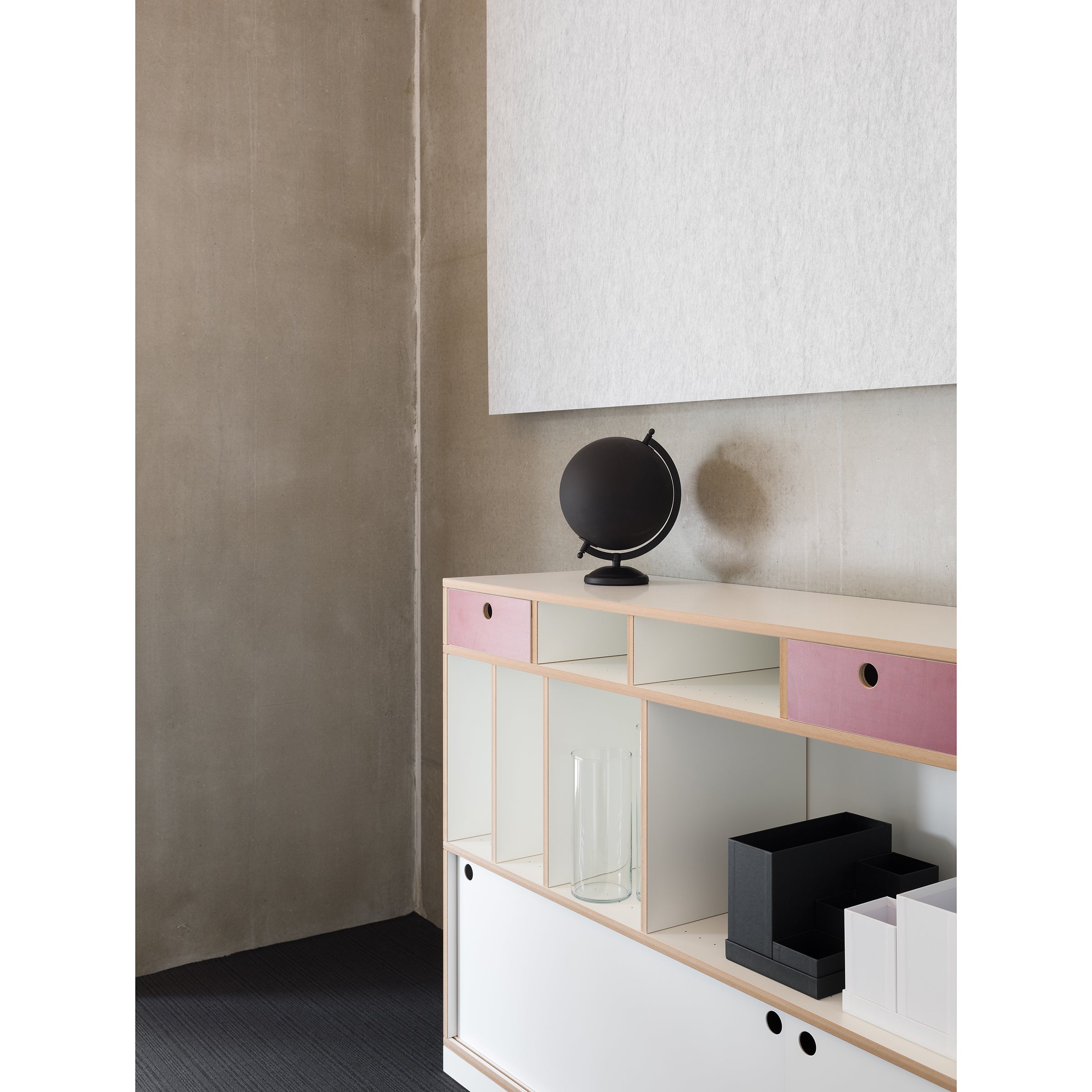 A minimalist room features the Moormann Egal cabinet with customizable wooden shelving, some highlighted by pink panels. Its adorned with a black globe and decorative items. Neutral walls display a large blank canvas above, contrasting beautifully with the dark floor.