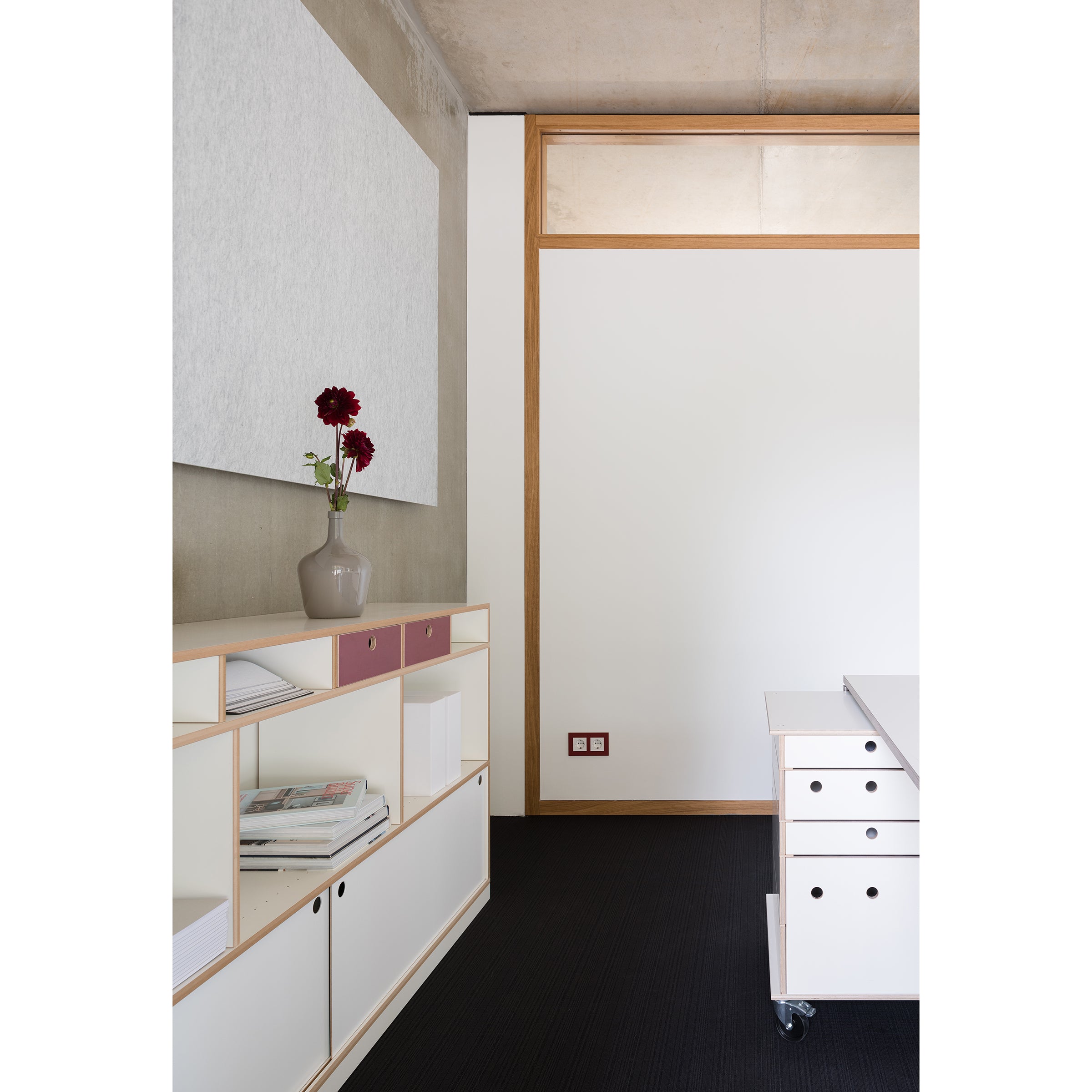 Minimalist office space with a white desk on wheels and an Egal shelf system by Moormann, featuring customizable shelving. A red flower vase adds color to the grey/white walls accented with wood, while a white panel decorates the wall above the versatile shelf unit.