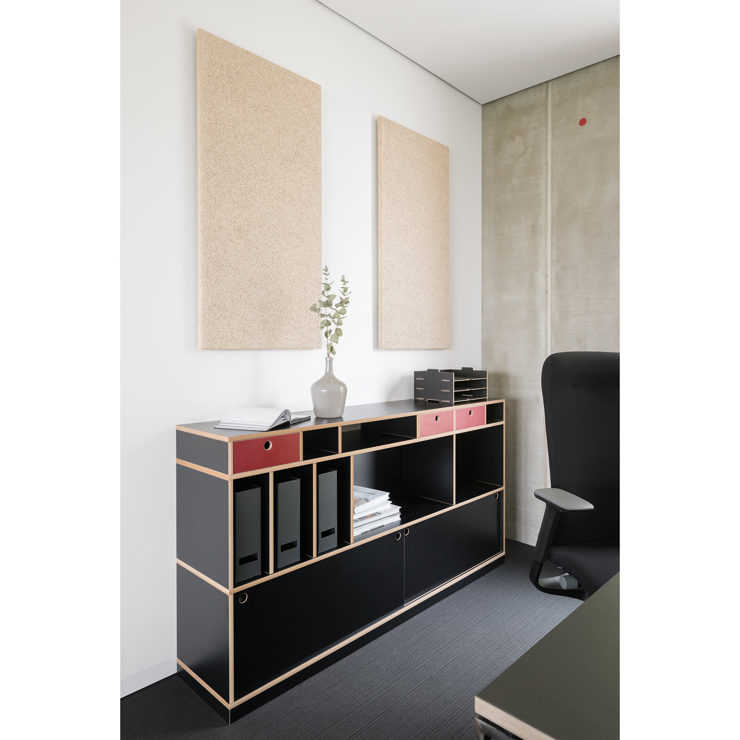 A modern office corner showcases the Moormann Egal shelf system with binders and decor, flanked by two acoustic panels on a white wall. On the right, a black office chair contrasts against a concrete wall.