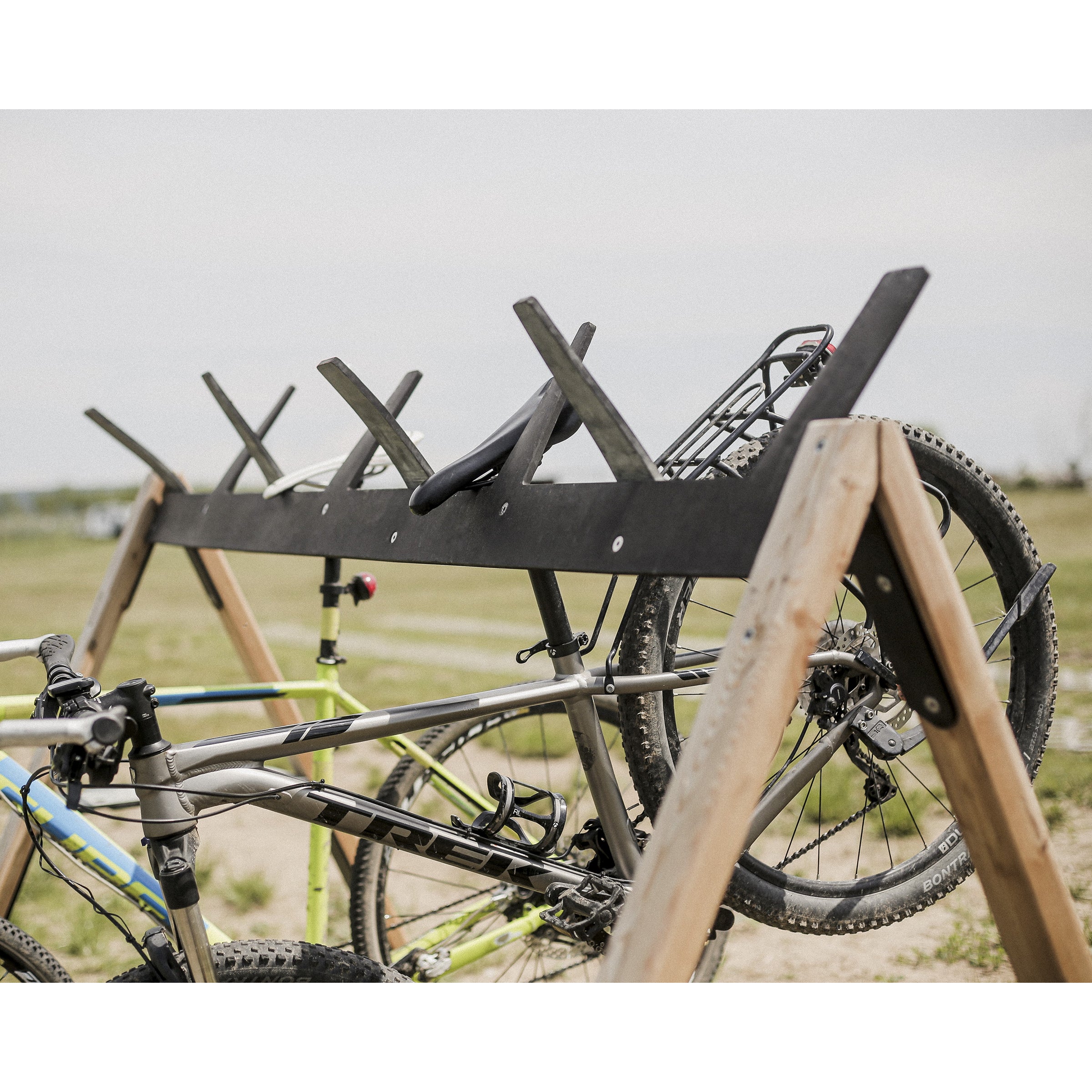 Two bicycles are placed on the multifunctional Cabra Ski and Bike Rack by Egoe, situated on grassy terrain. The wooden stand securely holds a gray bike and a light-colored one with visible handlebars and wheels, set against an overcast sky.