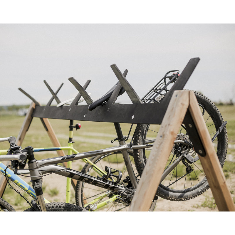 Two bicycles are placed on the multifunctional Cabra Ski and Bike Rack by Egoe, situated on grassy terrain. The wooden stand securely holds a gray bike and a light-colored one with visible handlebars and wheels, set against an overcast sky.