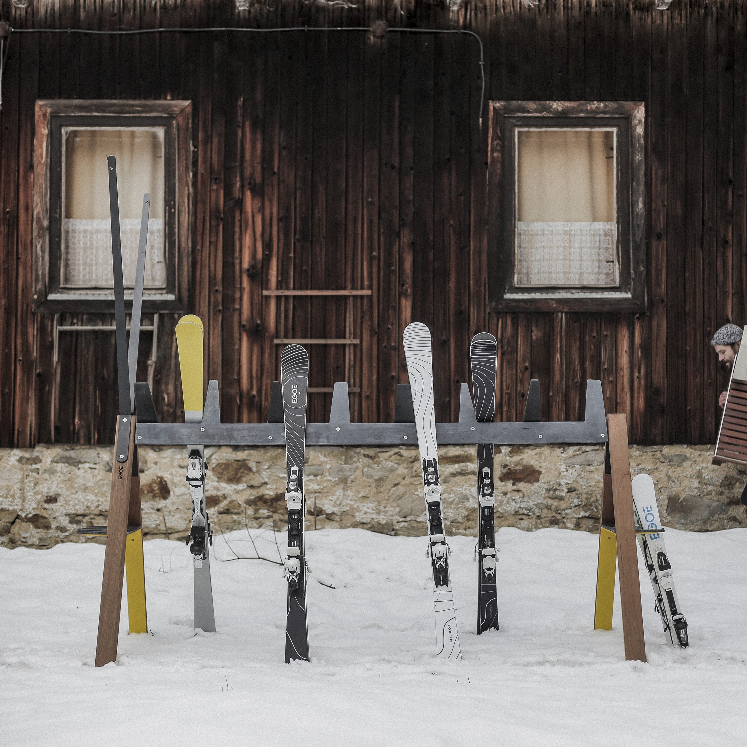 Skis are neatly arranged on the multifunctional Egoe Cabra Ski and Bike Rack in front of a rustic cabin with weathered wood and two curtained windows, while snow blankets the ground, creating a wintry scene.